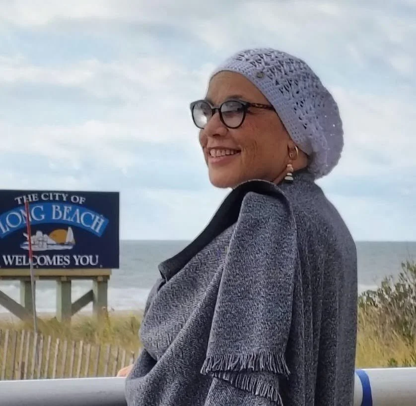 Woman smiling with glasses and earrings, wearing a gray coat and knit hat, standing outdoors near the beach, with a sign that reads "Long Beach welcomes you" in the background.