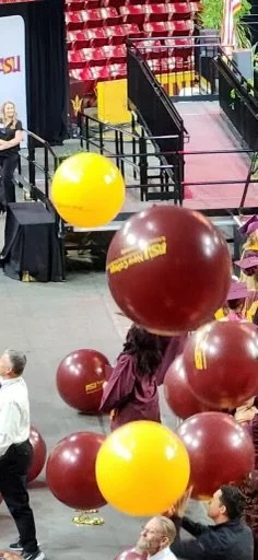People holding large yellow and maroon balloons at an indoor event with a stage and stairs in the background.