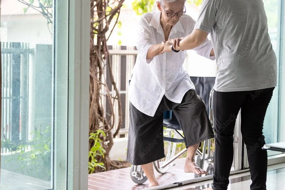 A caregiver assisting an elderly woman with a mobility aid indoors, holding her arm as she steps outside through a glass door.