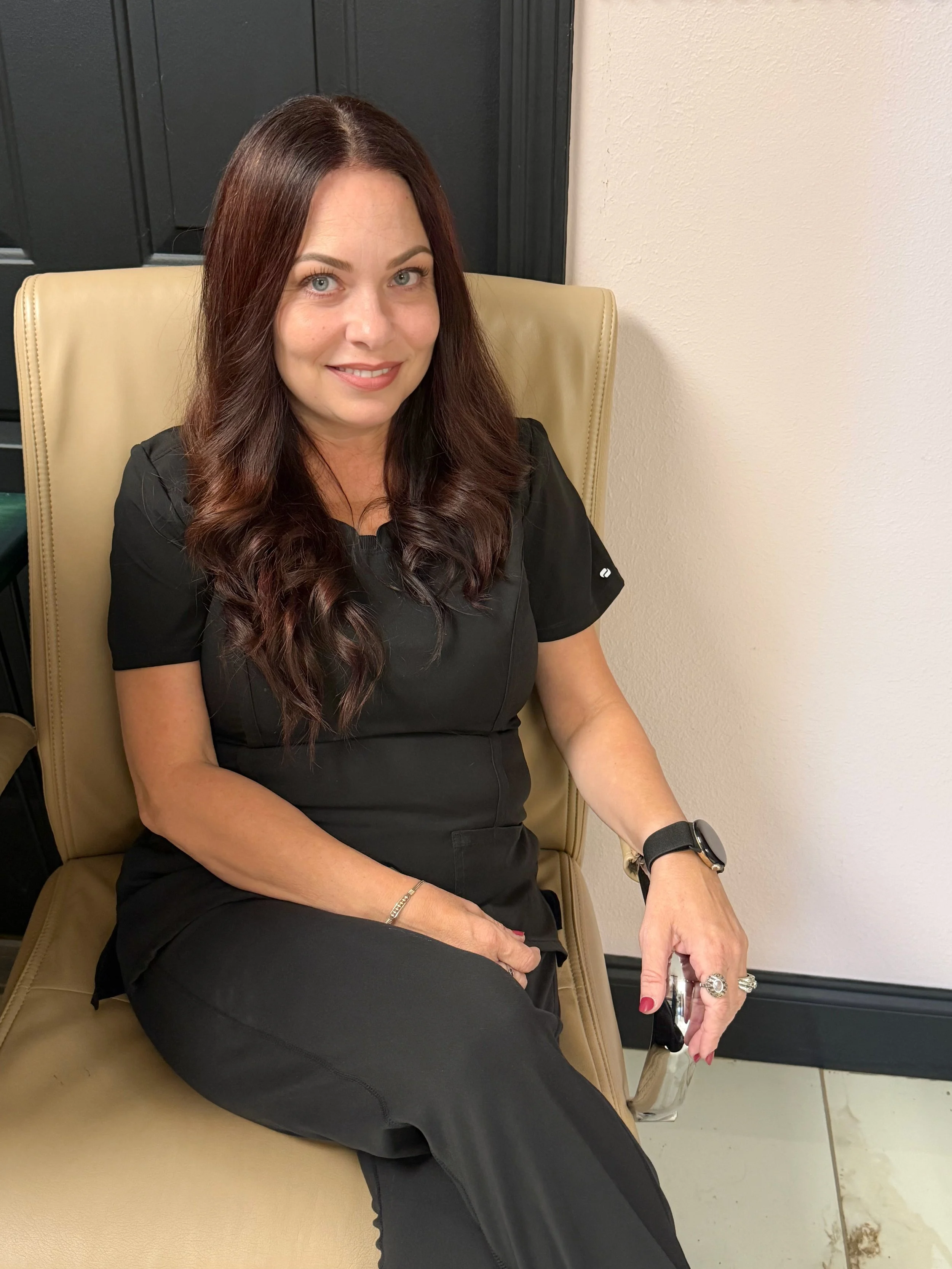 A woman with long brown hair, light skin, and blue eyes sitting in a beige chair. She is wearing a black short-sleeved top, a watch, large rings, and has painted nails. She is smiling softly and looking at the camera, sitting with her arm resting on her lap and Her right hand holding a smartphone.