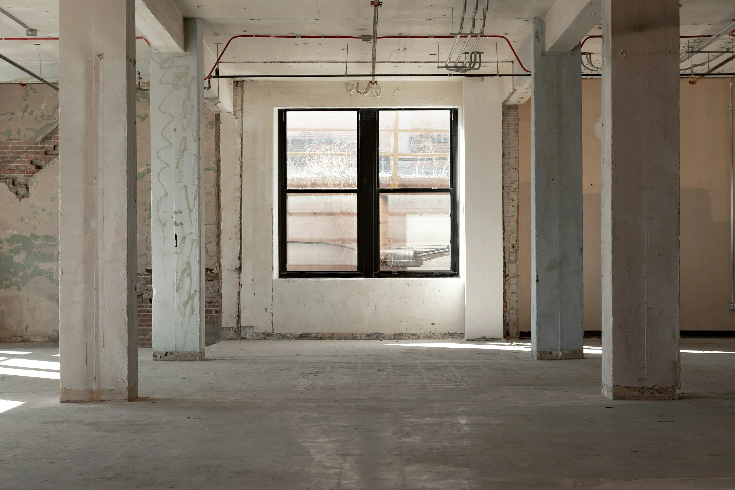 Interior of a building under construction with exposed concrete columns, brick walls, and a window letting in natural light.