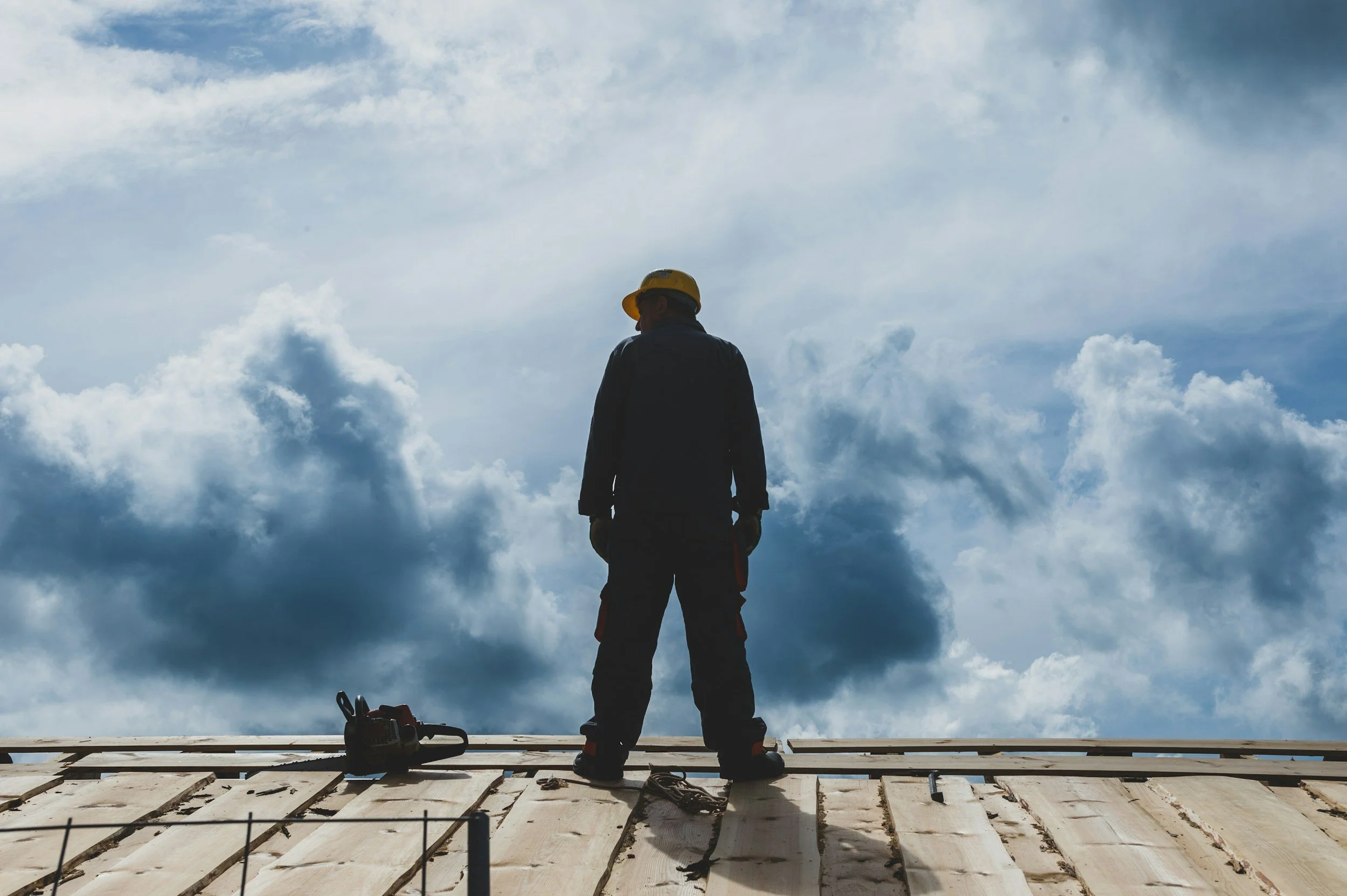 Construction worker wearing a yellow helmet standing on a wooden platform with tools nearby, against a cloudy sky.