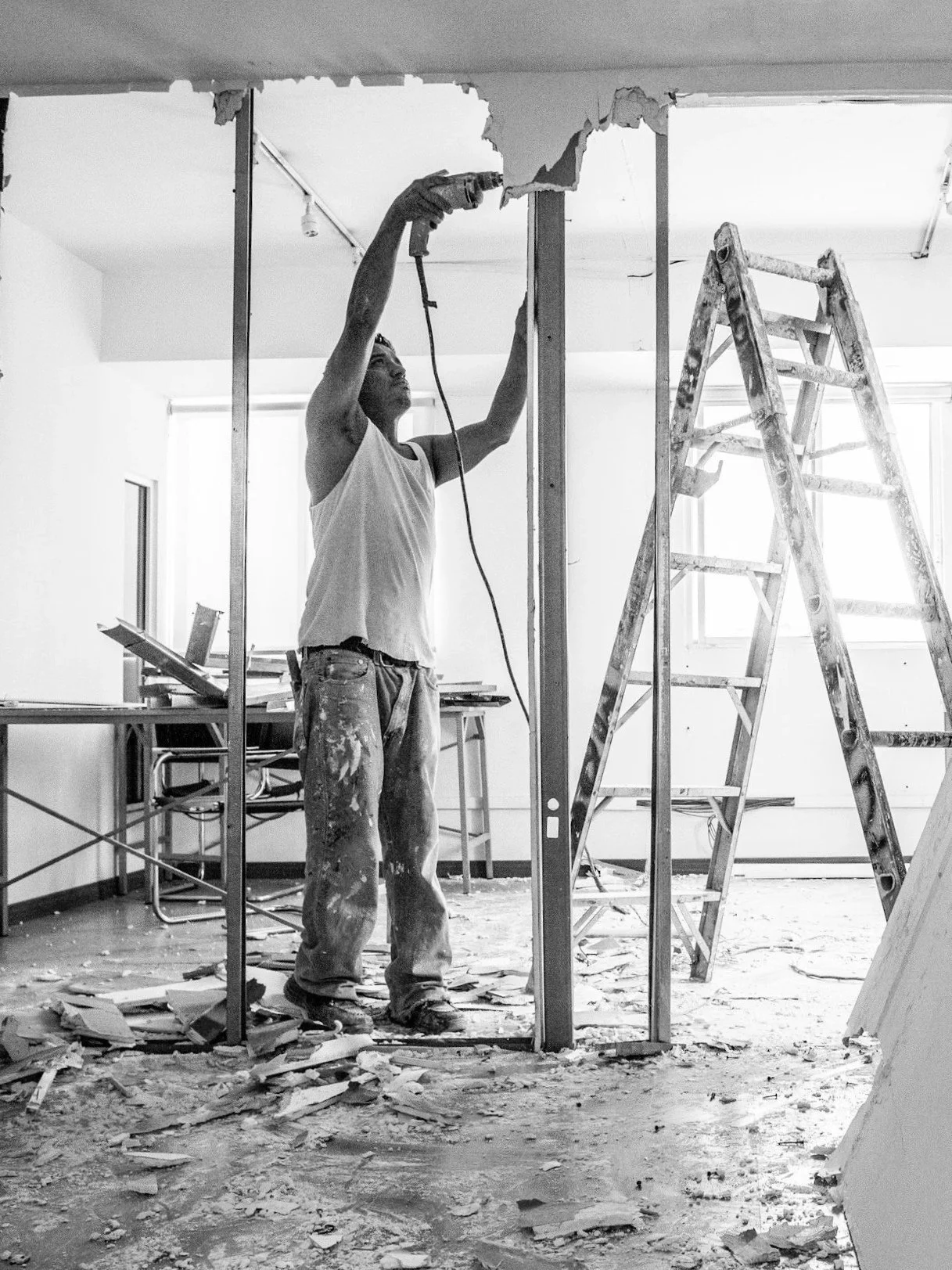 A man working on construction inside a house, using a power drill on a ceiling. Construction materials and a ladder are visible in the background.