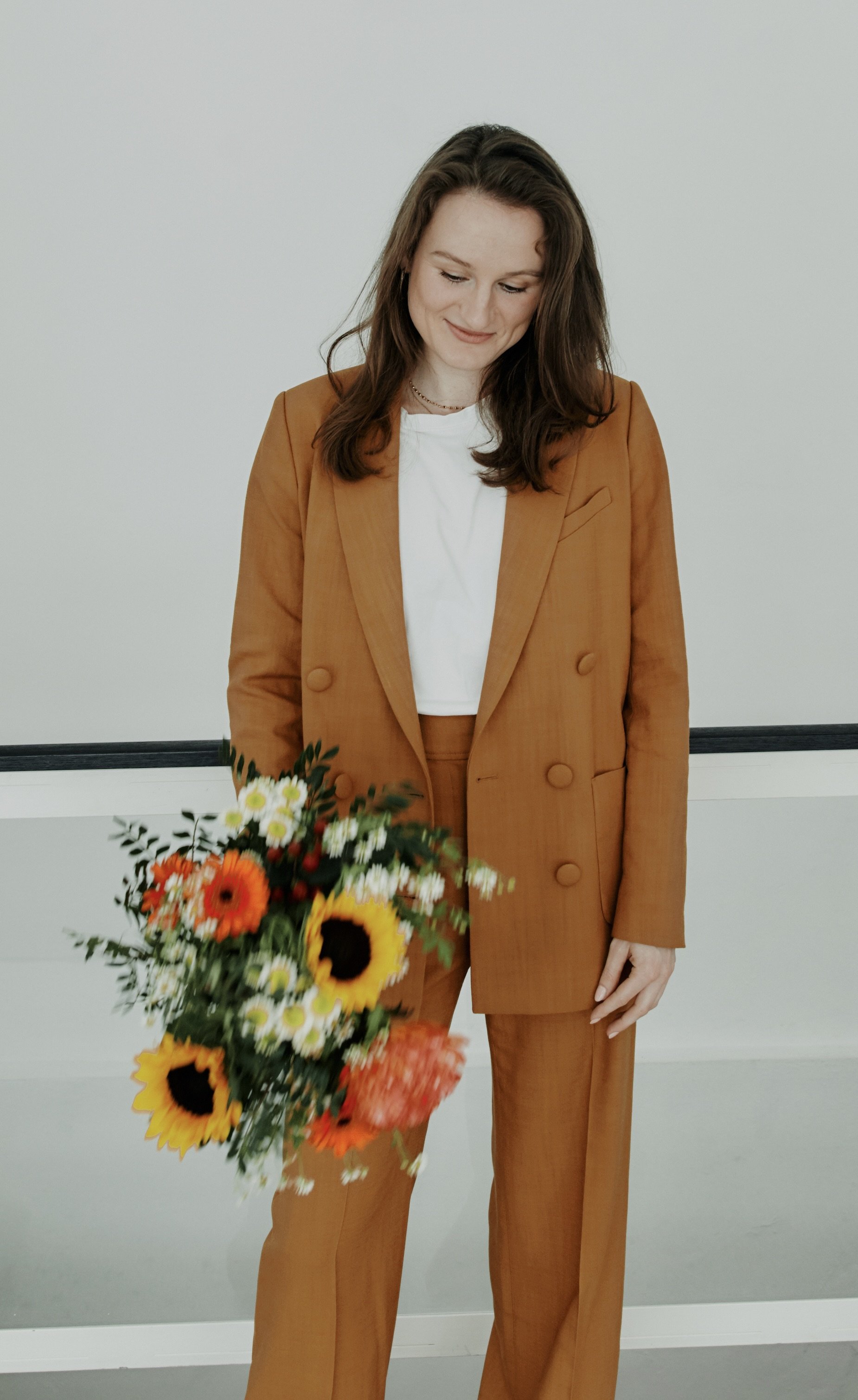 A young woman with brown hair wearing an orange suit and white shirt, standing against a plain wall, holding a colorful bouquet of flowers including sunflowers, daisies, and orange blooms.