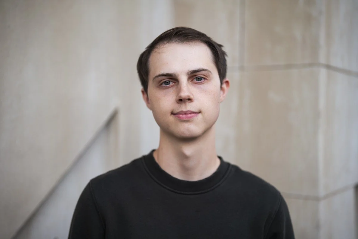A young man with short dark hair, wearing a black shirt, standing in front of a neutral-colored wall.