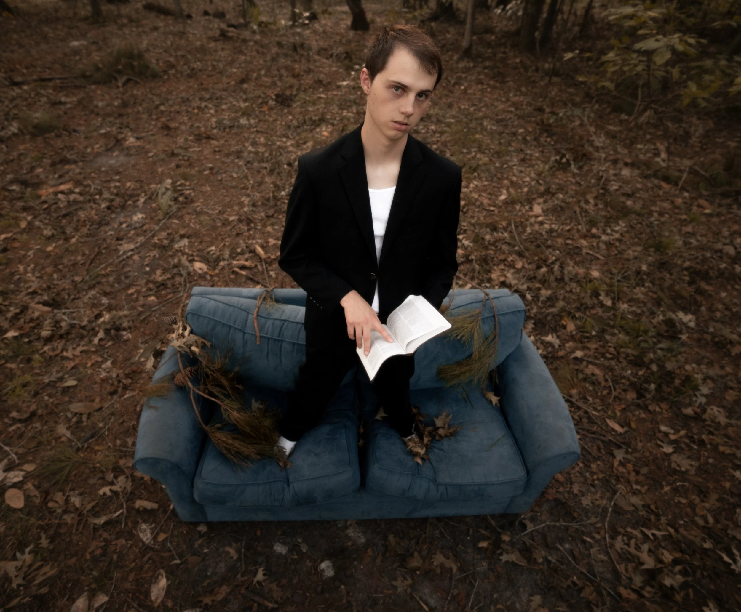 A young man with short dark hair and wearing a black blazer over a white shirt sits on a blue cushioned bench outdoors in a forested area with brown fallen leaves. He is holding an open book.