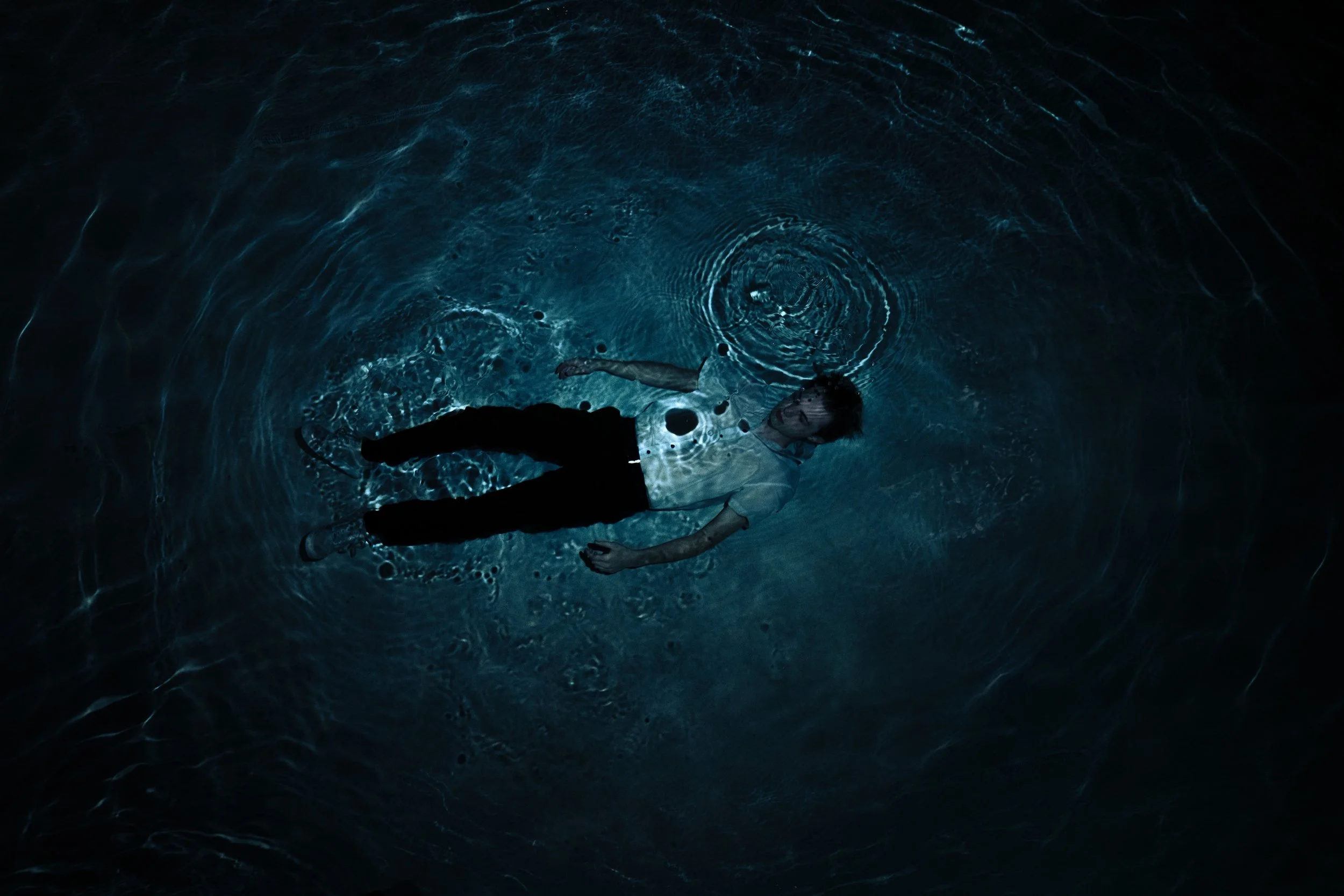 A person lying on their back in a dark water pool, illuminated by subtle blue light reflecting off the water's surface.