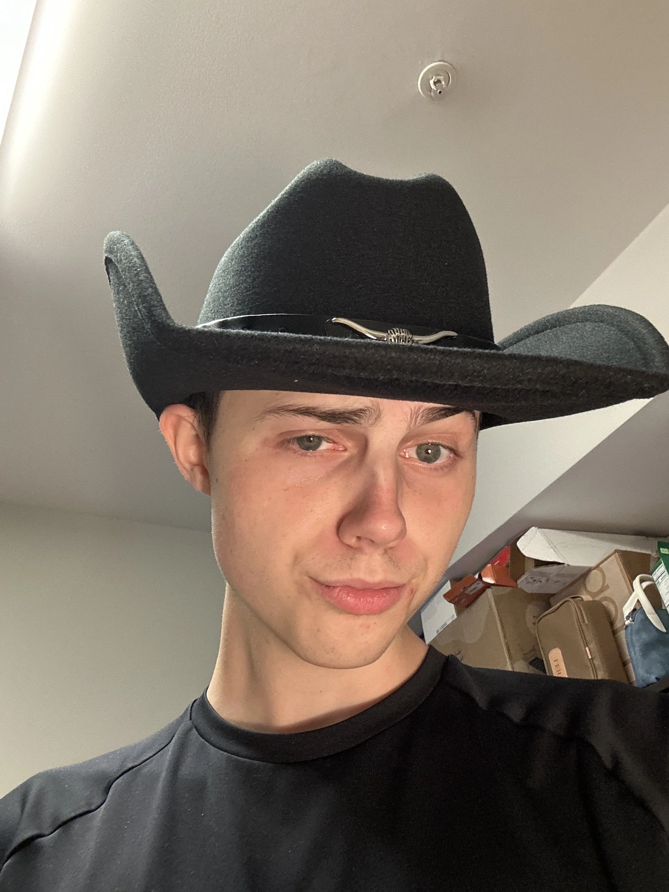 Person wearing a black cowboy hat with a metal band, standing indoors with boxes on shelves in the background.