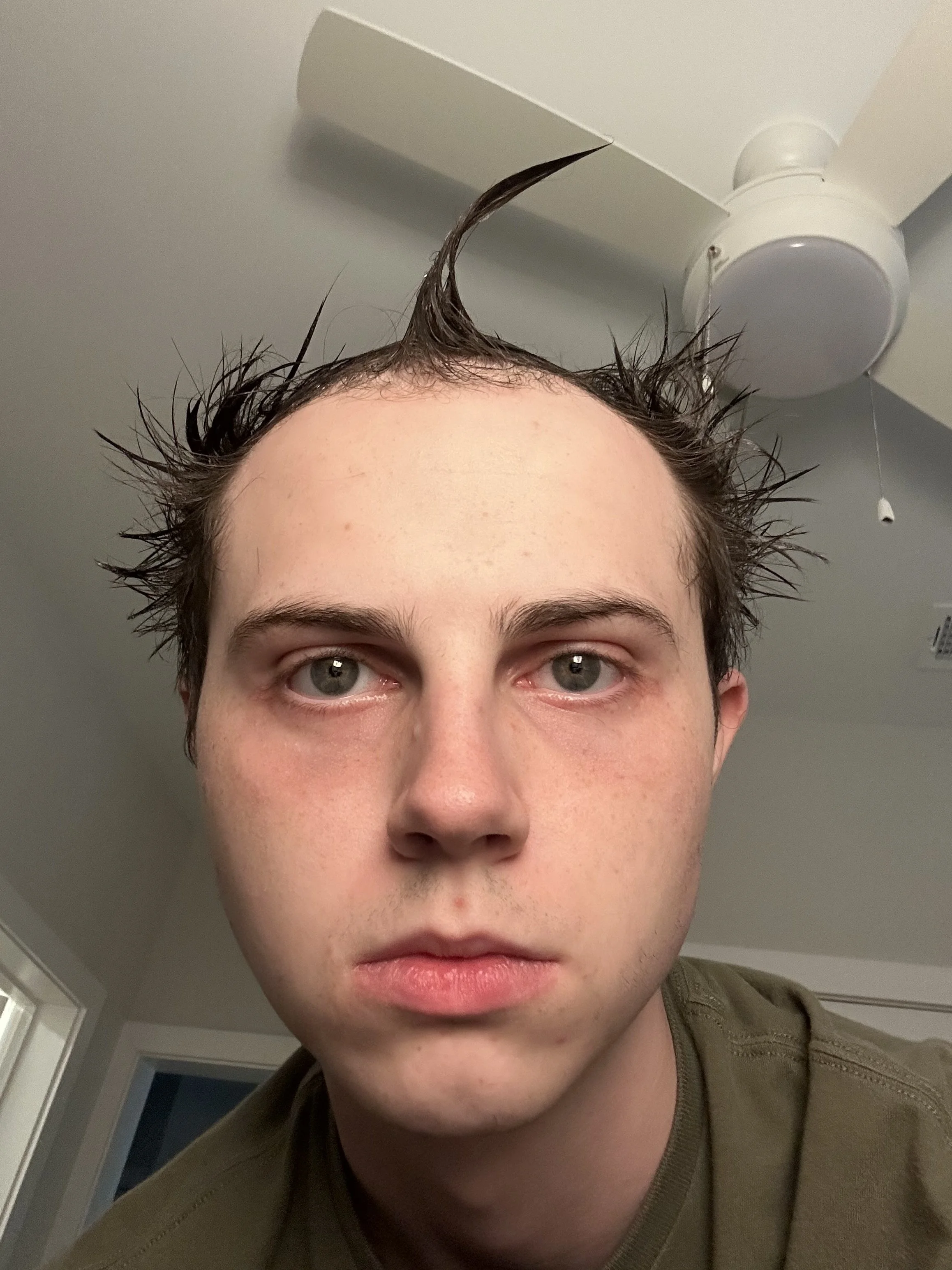 Close-up of a young person with damp, spiky hair looking directly at the camera, with part of their face visible, in a room with a ceiling fan in the background.