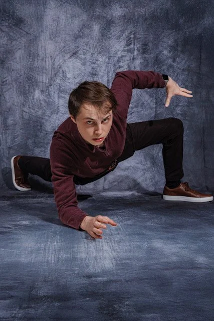 A young man in a maroon sweater and black pants is in a crouched martial arts pose on the ground, against a textured blue-gray background.