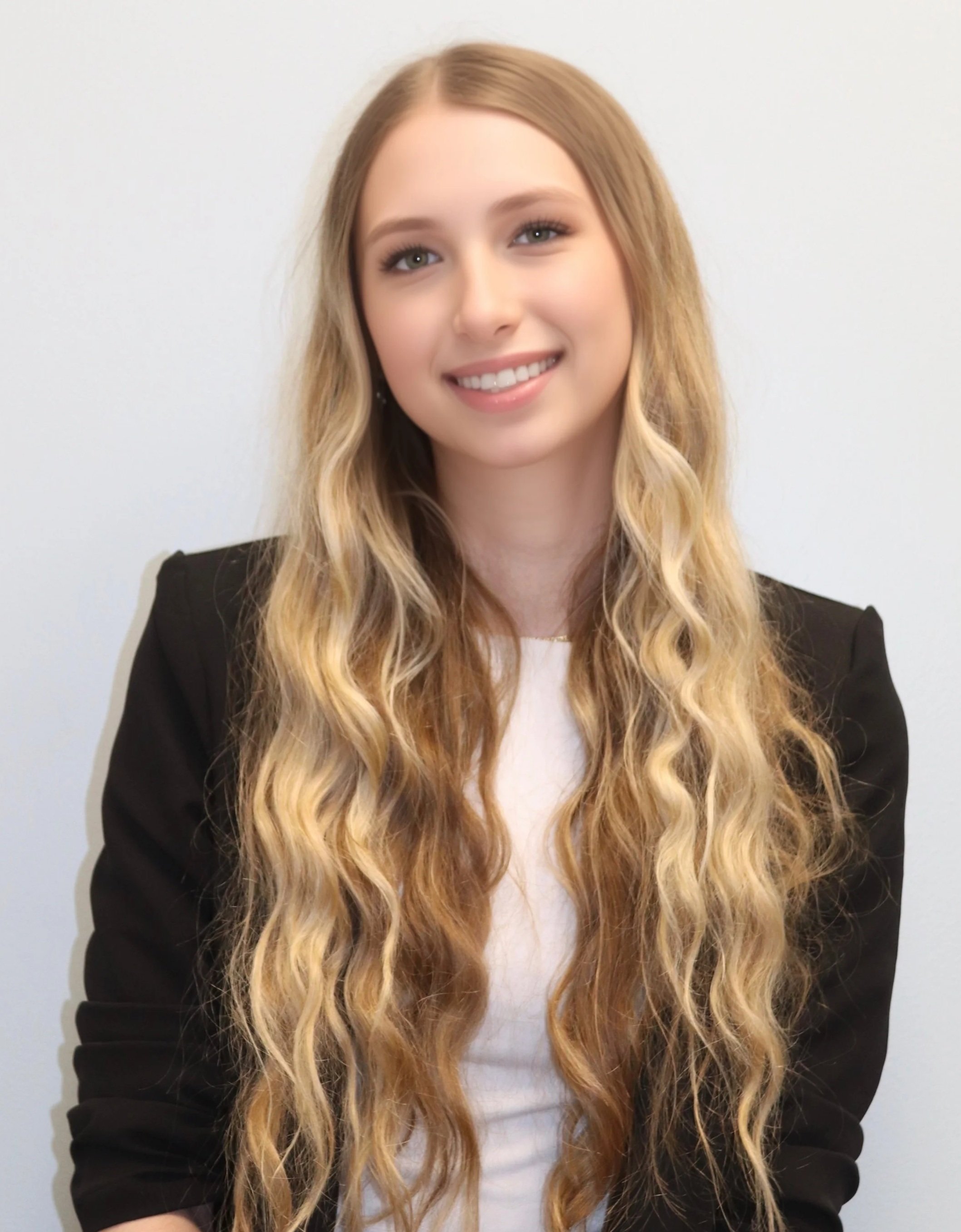 Daniela, a hearing specialist, with long wavy blonde hair smiling at the camera, wearing a black blazer over a white top, standing against a plain white wall.