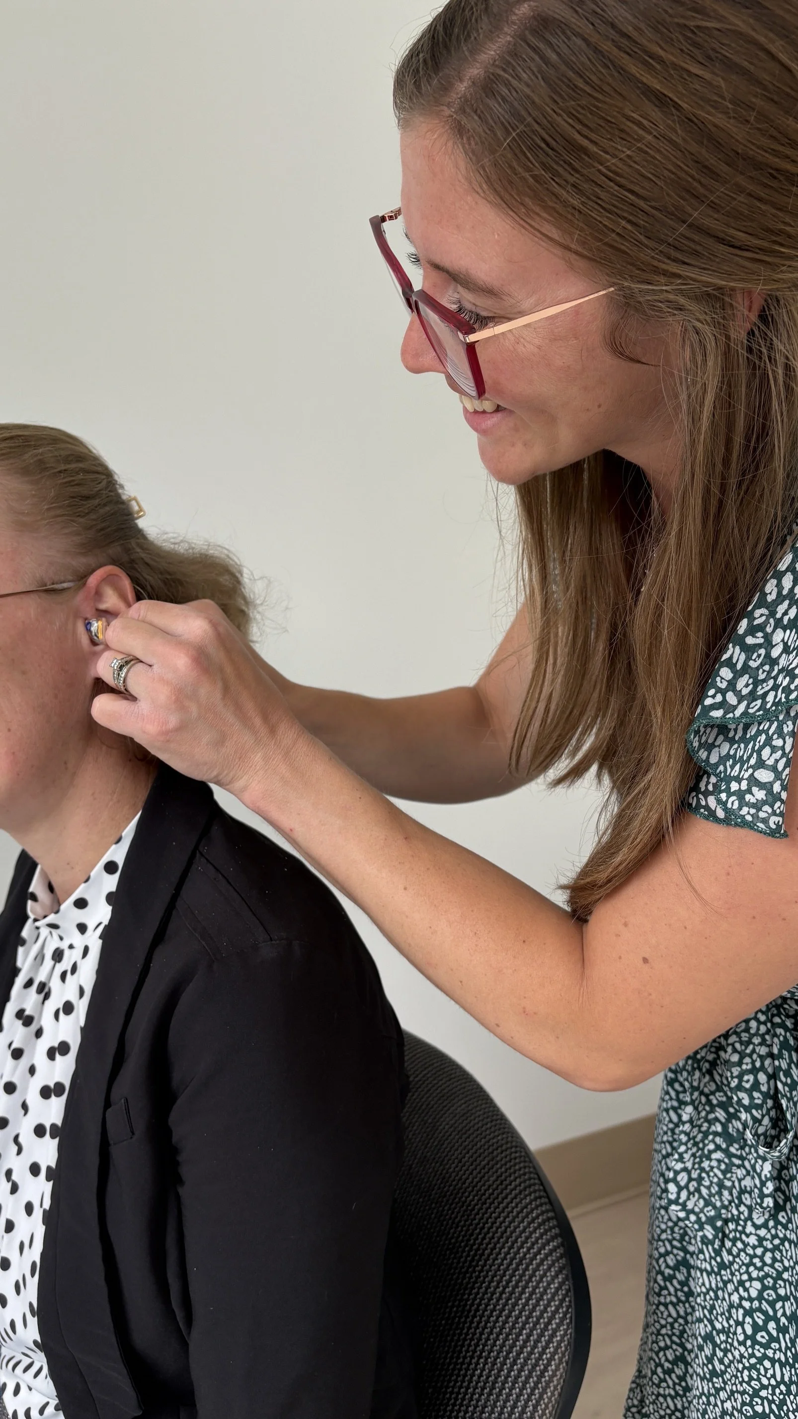 A woman with long brown hair and glasses is examining a woman with curly red hair, wearing a black jacket and a polka dot blouse, who is sitting in a chair.