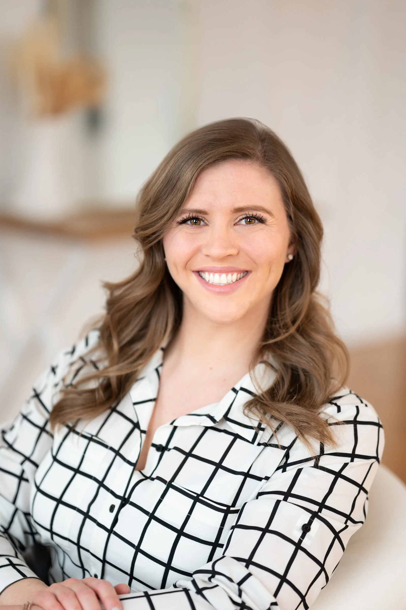 Becca, business owner and hearing specialist, with light brown hair styled in loose waves, wearing a white blouse with a black grid pattern, smiling brightly with white teeth, seated in a well-lit room with a neutral background.