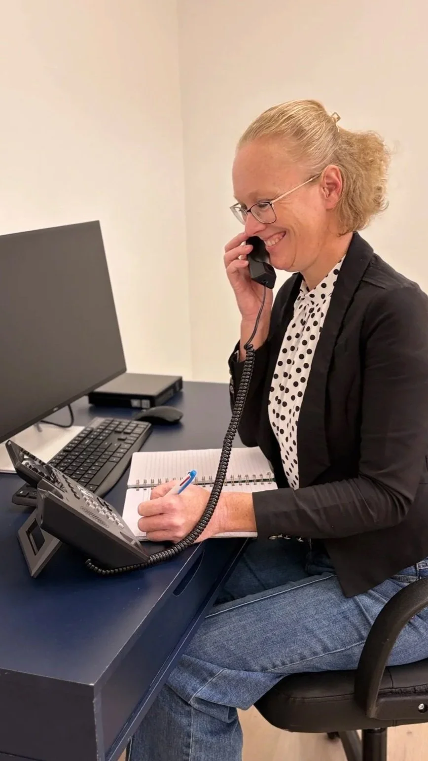 Woman sitting at a desk, talking on a landline phone, with a notepad and pen, computer monitor, keyboard, and mouse on the desk.