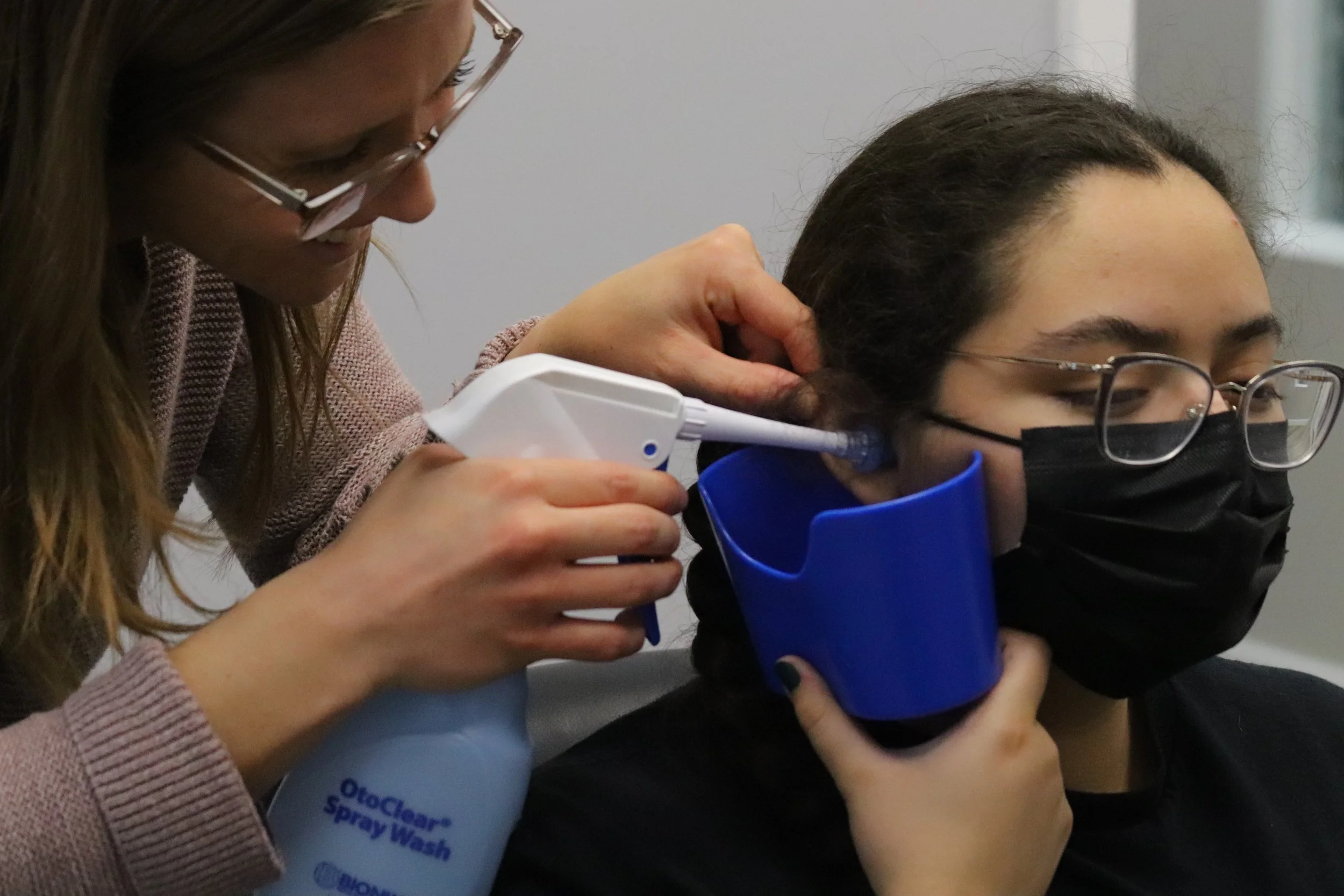 A hearing specialist is looking at the ear of another young woman who is wearing glasses and a black face mask.