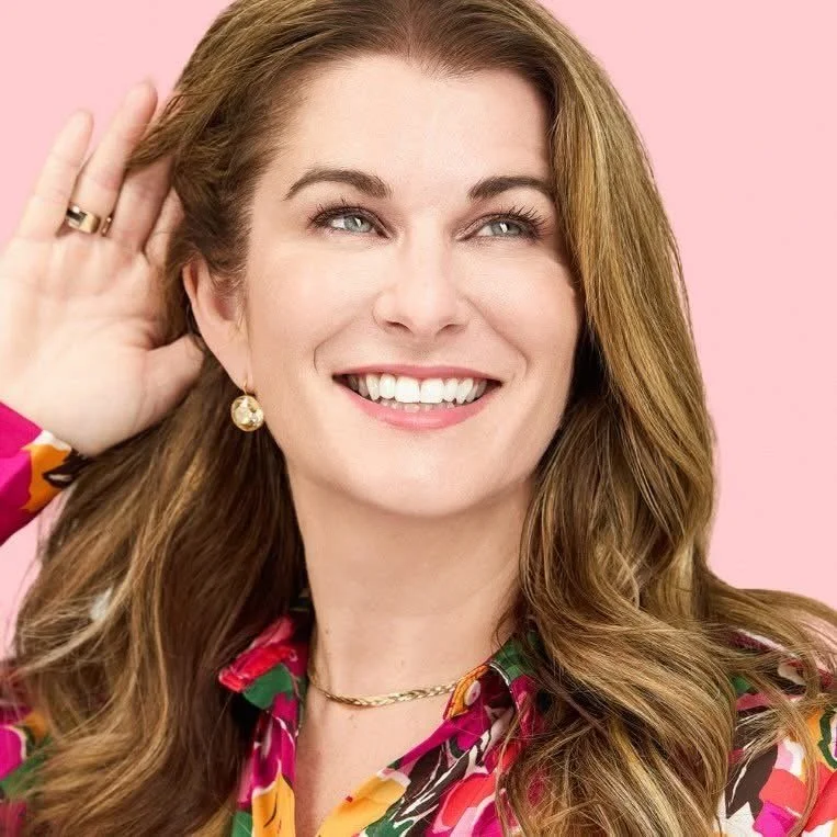 A smiling woman with long, wavy brown hair, wearing a colorful floral blouse, gold earrings, and a gold necklace, posed against a pink background.