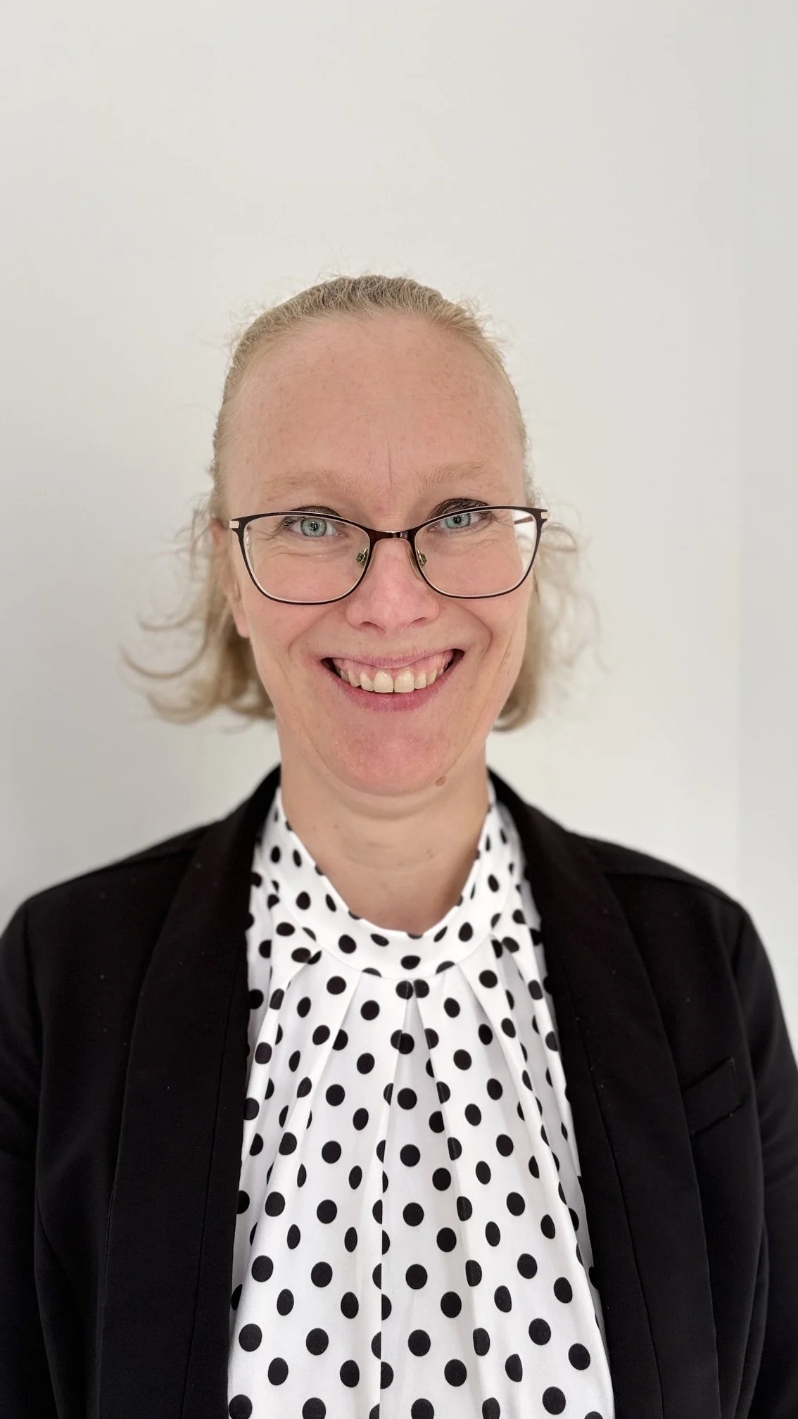 Smiling woman with glasses, blonde curly hair pulled back, wearing a black blazer over a white blouse with black polka dots, in front of a plain white background.