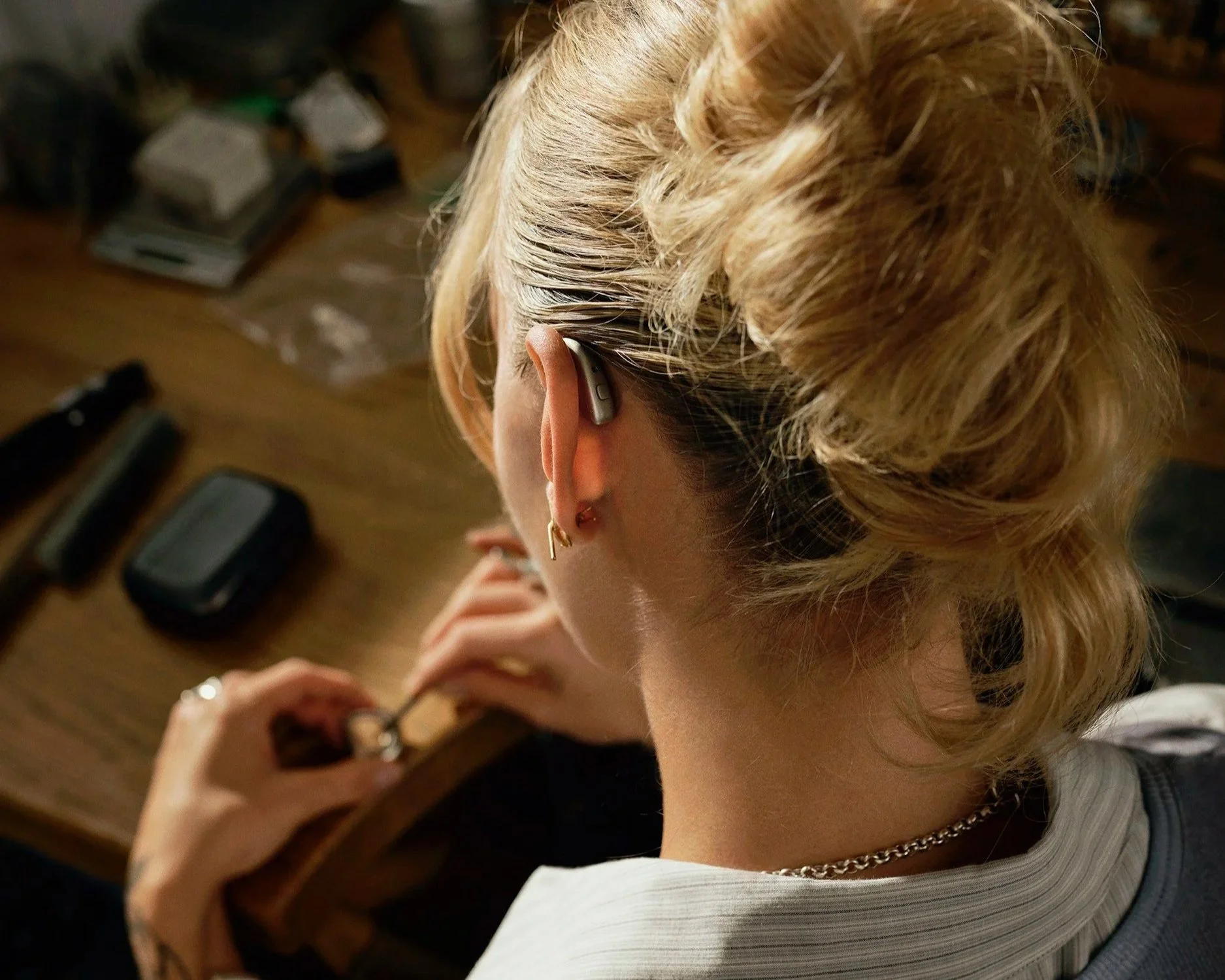 A person with blonde hair wearing a hearing aid. The background includes a wooden surface with electronic devices and cables.