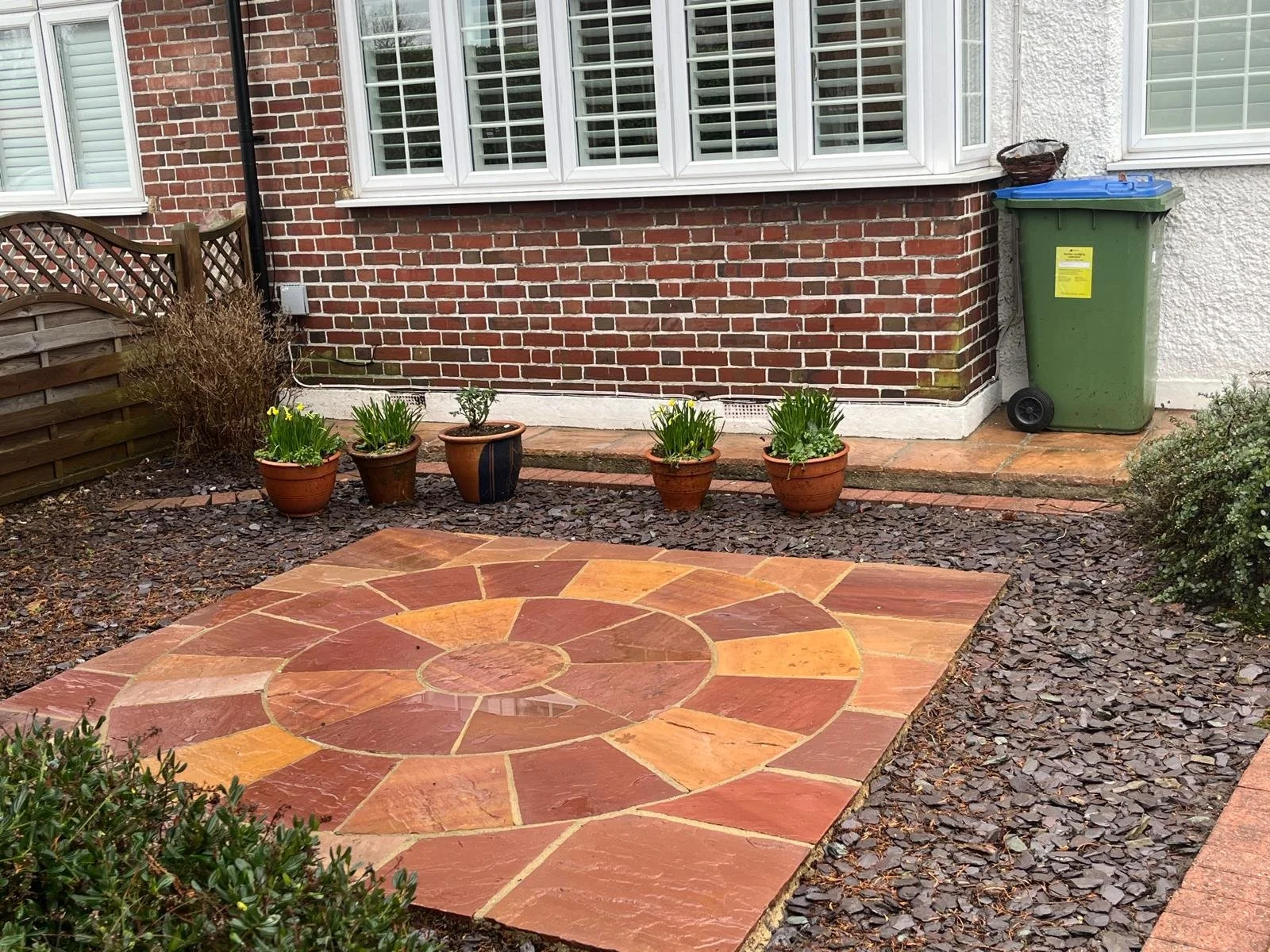 Backyard patio with a circular pattern of red and yellow stone tiles, potted plants in front of a brick house wall, and a green trash bin with a blue lid on the right side.