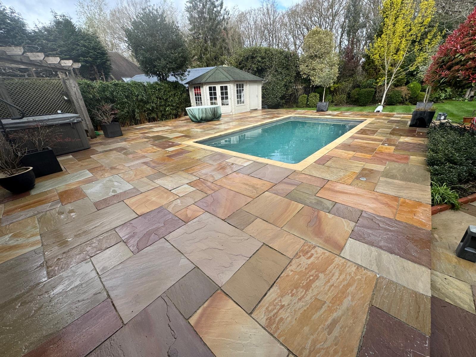 Backyard with a pool, surrounded by multi-colored stone pavers, trees, and shrubs.