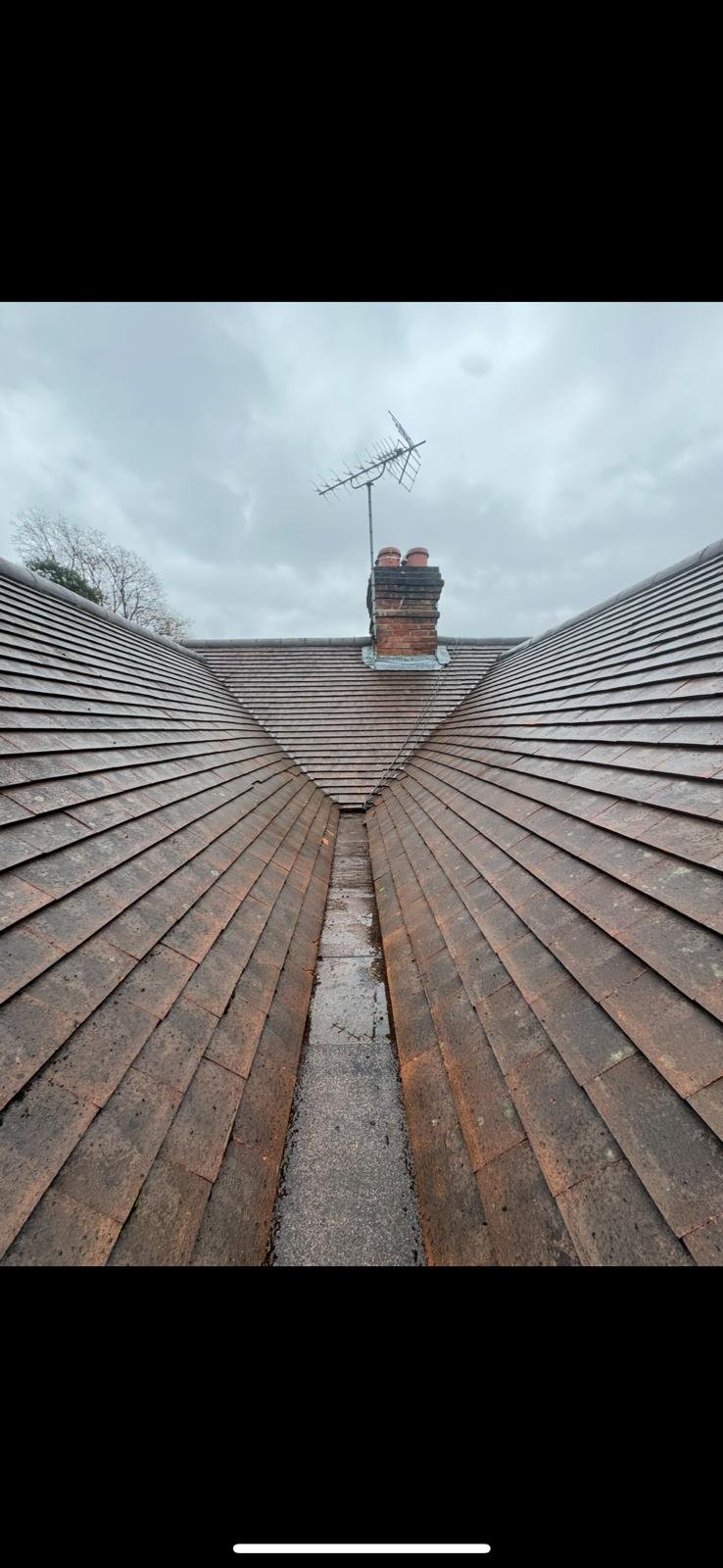 View of a roof with brown shingles and a central chimney with an old-fashioned television antenna on top, under a cloudy sky.