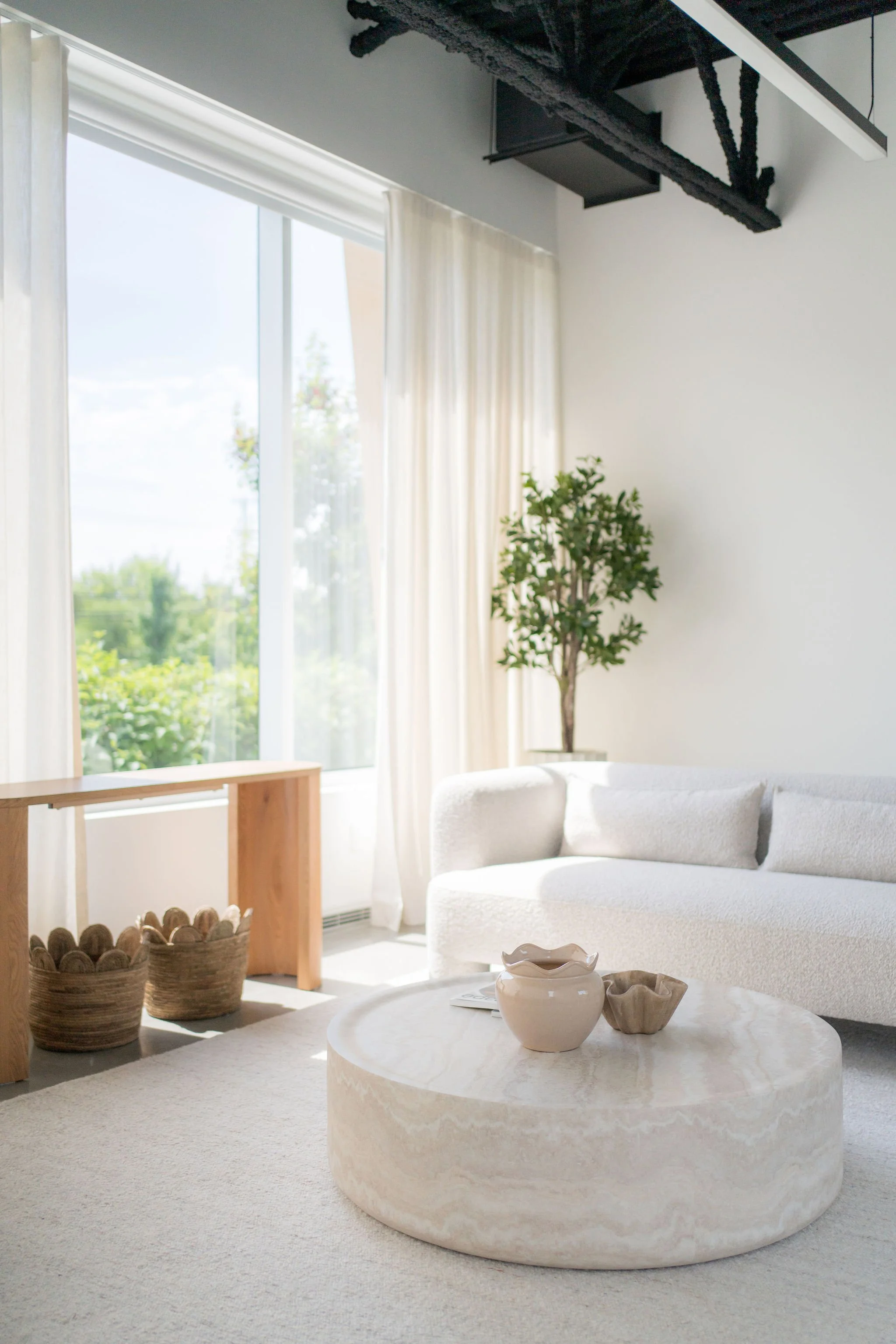 Salon lumineux avec grande fenêtre, canapé blanc, table basse en pierre, décor minimaliste avec potées en osier, plantes vertes, et décorations en céramique.