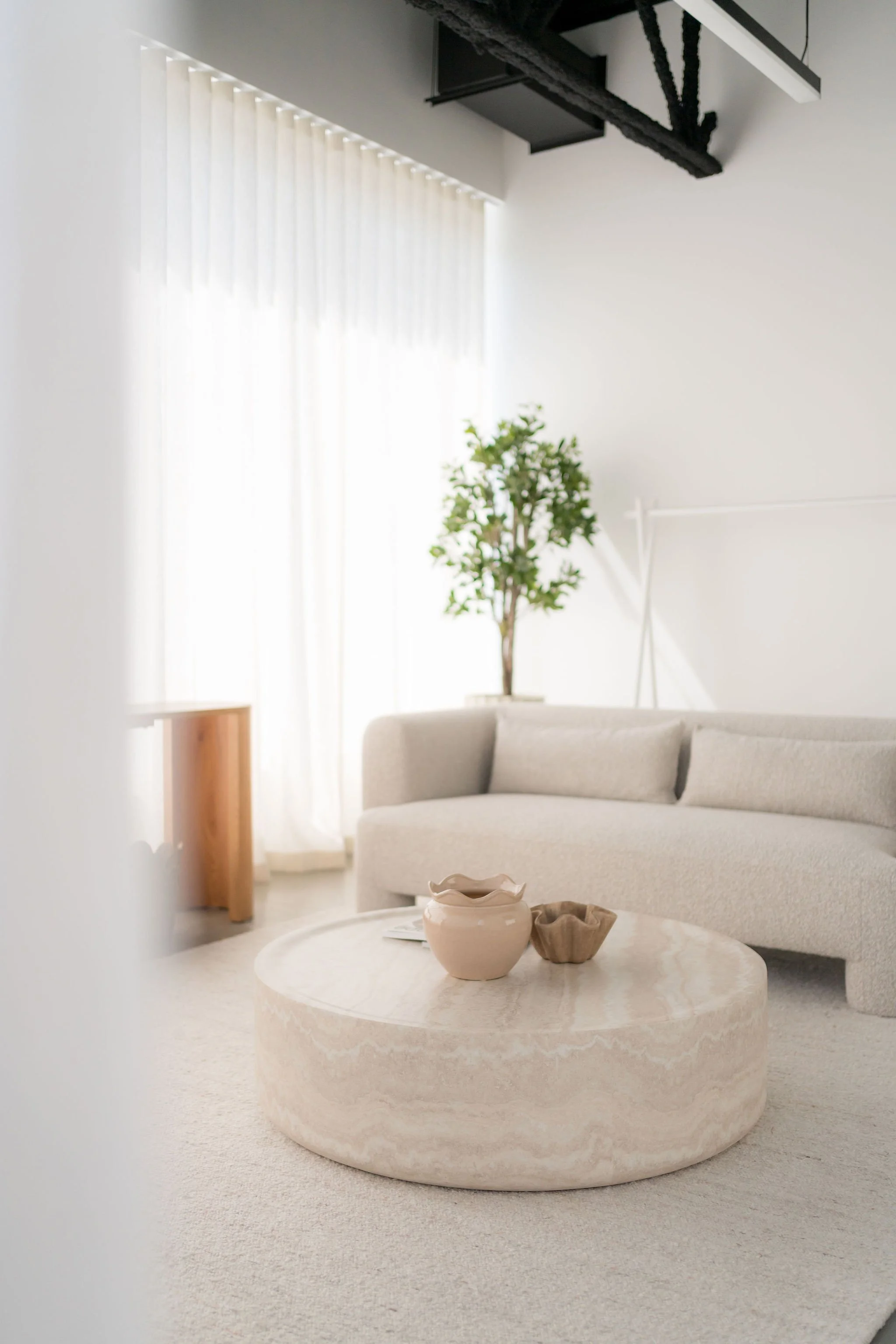 Salon lumineux avec un canapé beige, une table ronde en pierre avec des vases en céramique, et une plante verte en arrière-plan. Lumière naturelle venant de grandes fenêtres avec des rideaux blancs.