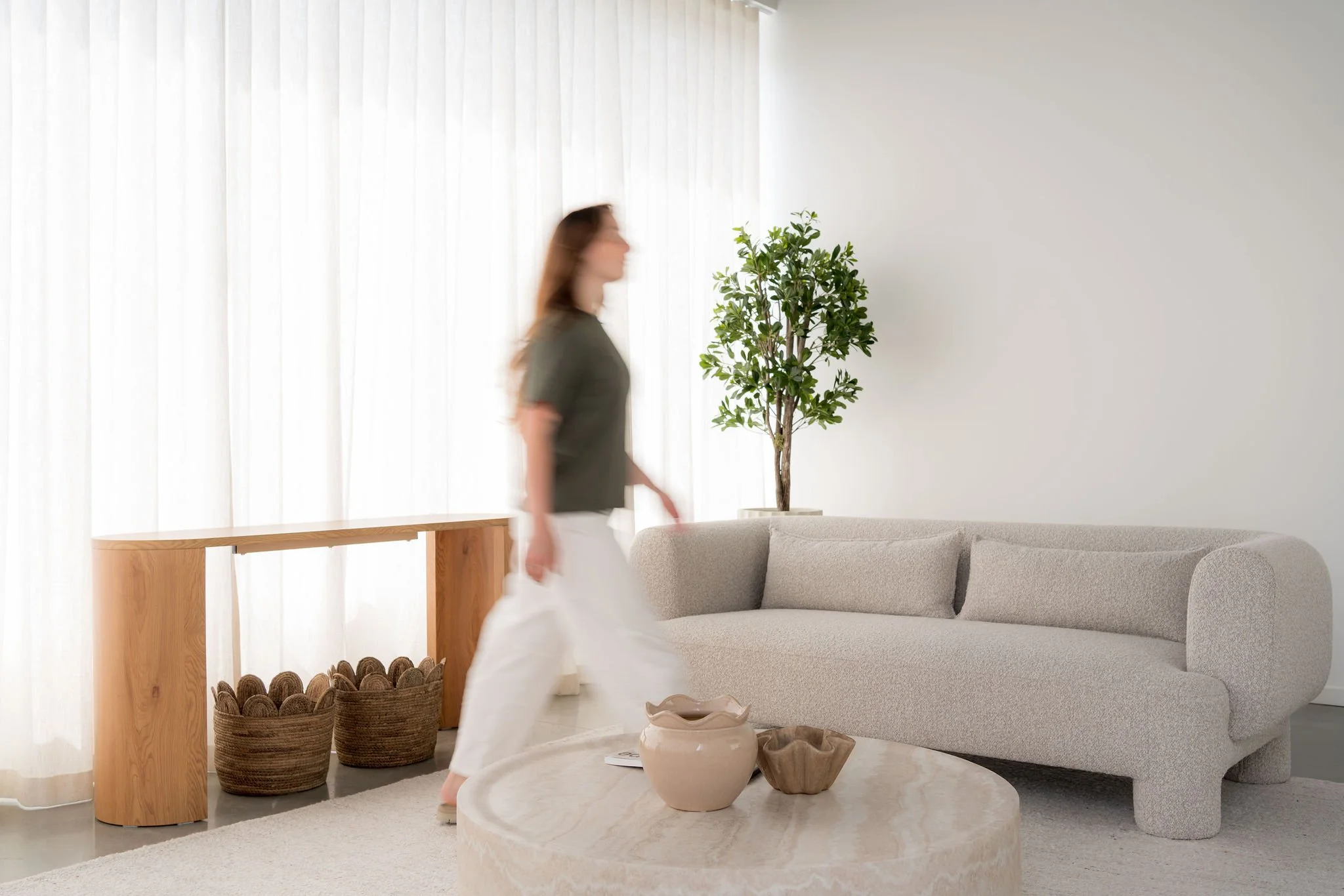 Une femme marche dans un salon moderne avec un sofa gris, une table en marbre, plusieurs paniers en osier, et une plante verte dans un pot, avec des rideaux blancs en arrière-plan.