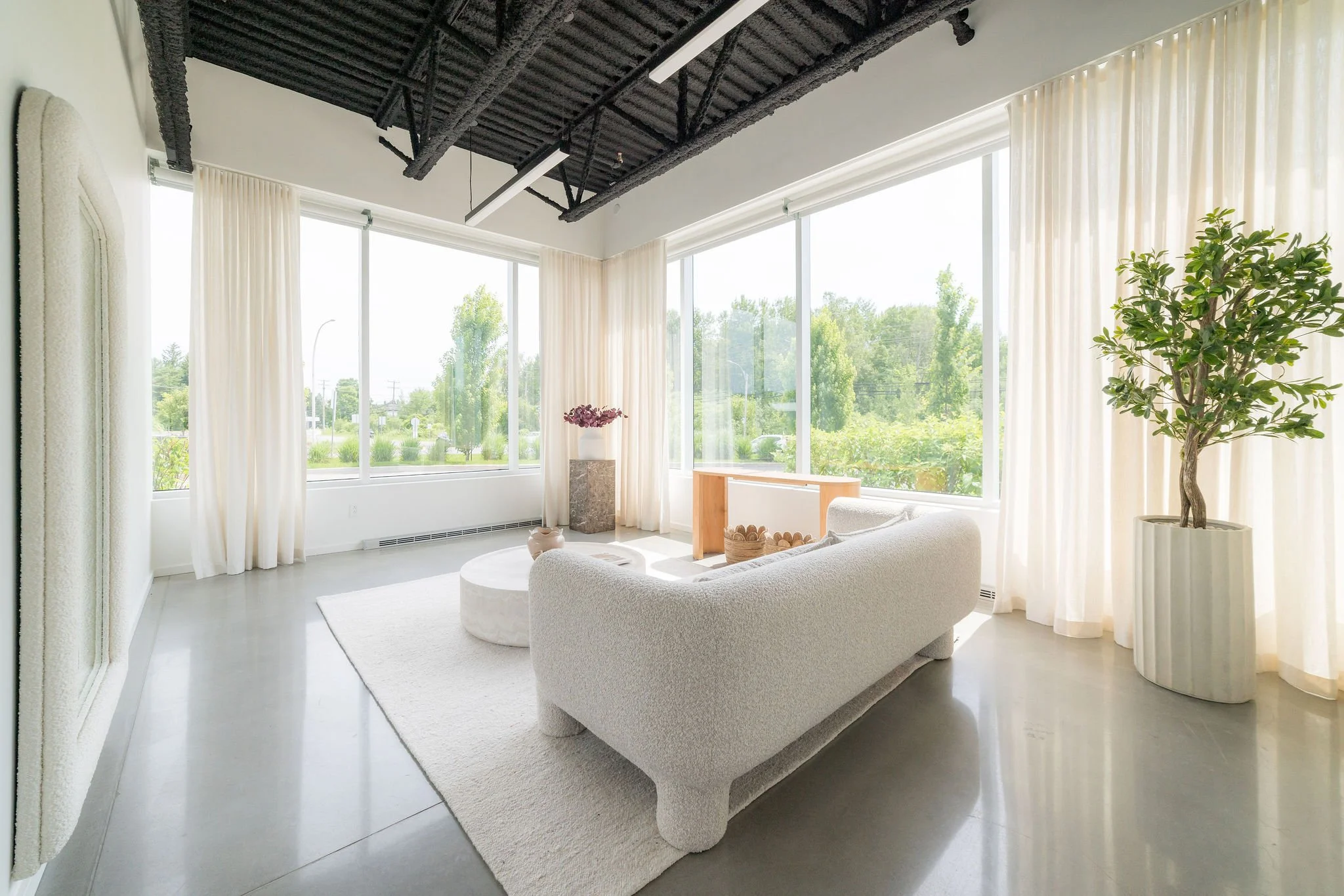 Salon moderne avec canapé blanc, grande baie vitrée, rideaux blancs, plante verte, table en bois, décoration minimaliste, vue de l'extérieur avec arbres et ciel clair.