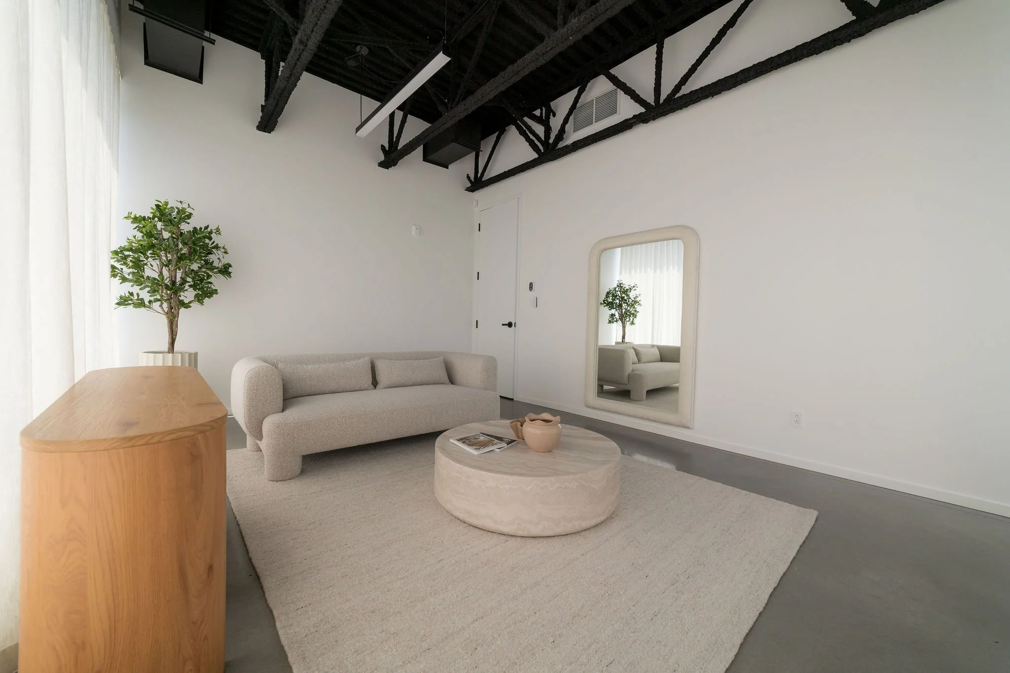 Un salon moderne avec un canapé beige, une table en marbre, un miroir rectangulaire, une plante verte, et un plafond avec poutres noires exposées.