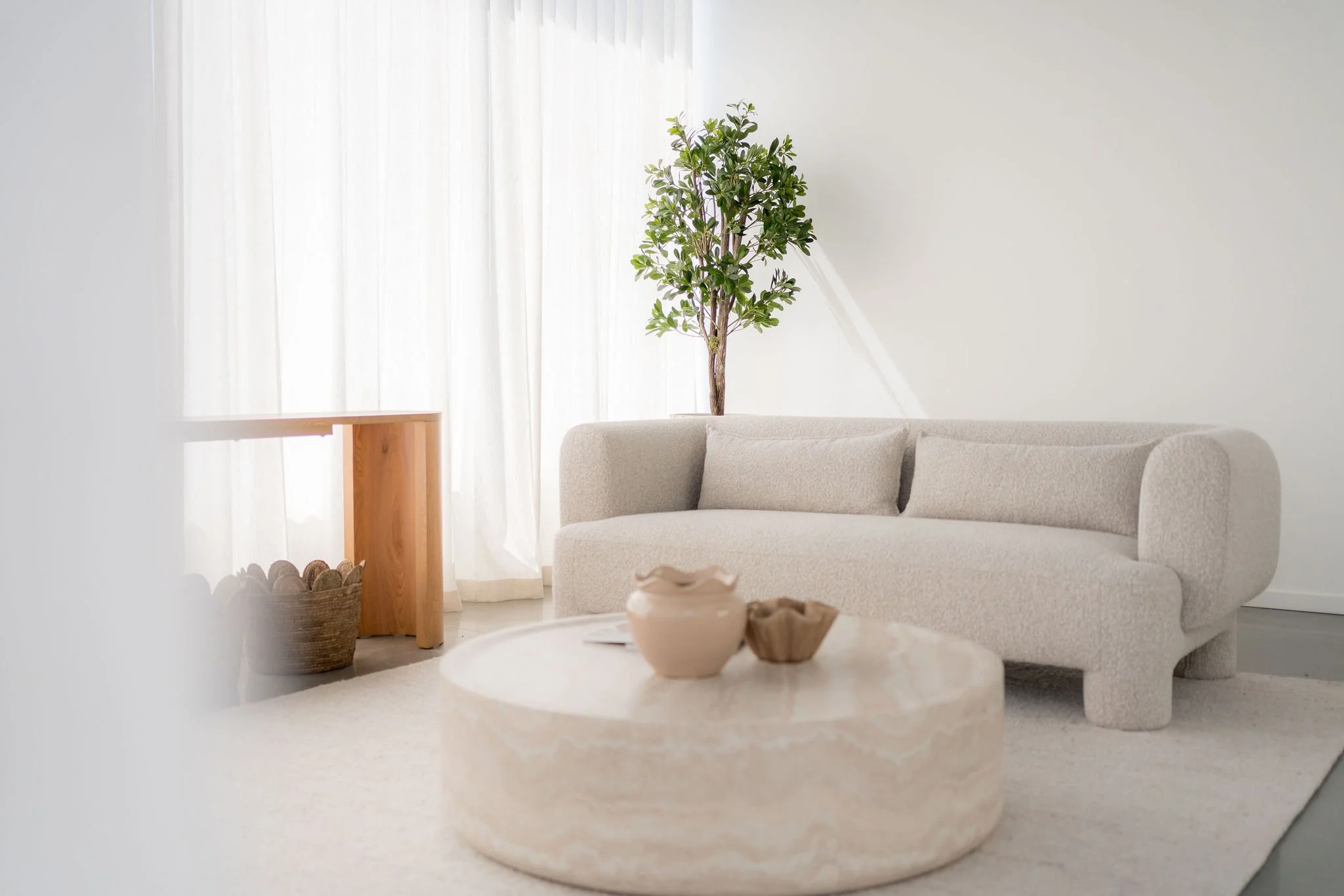 Salon minimaliste avec canapé beige, table basse en pierre, plantes vertes et rideaux blancs.