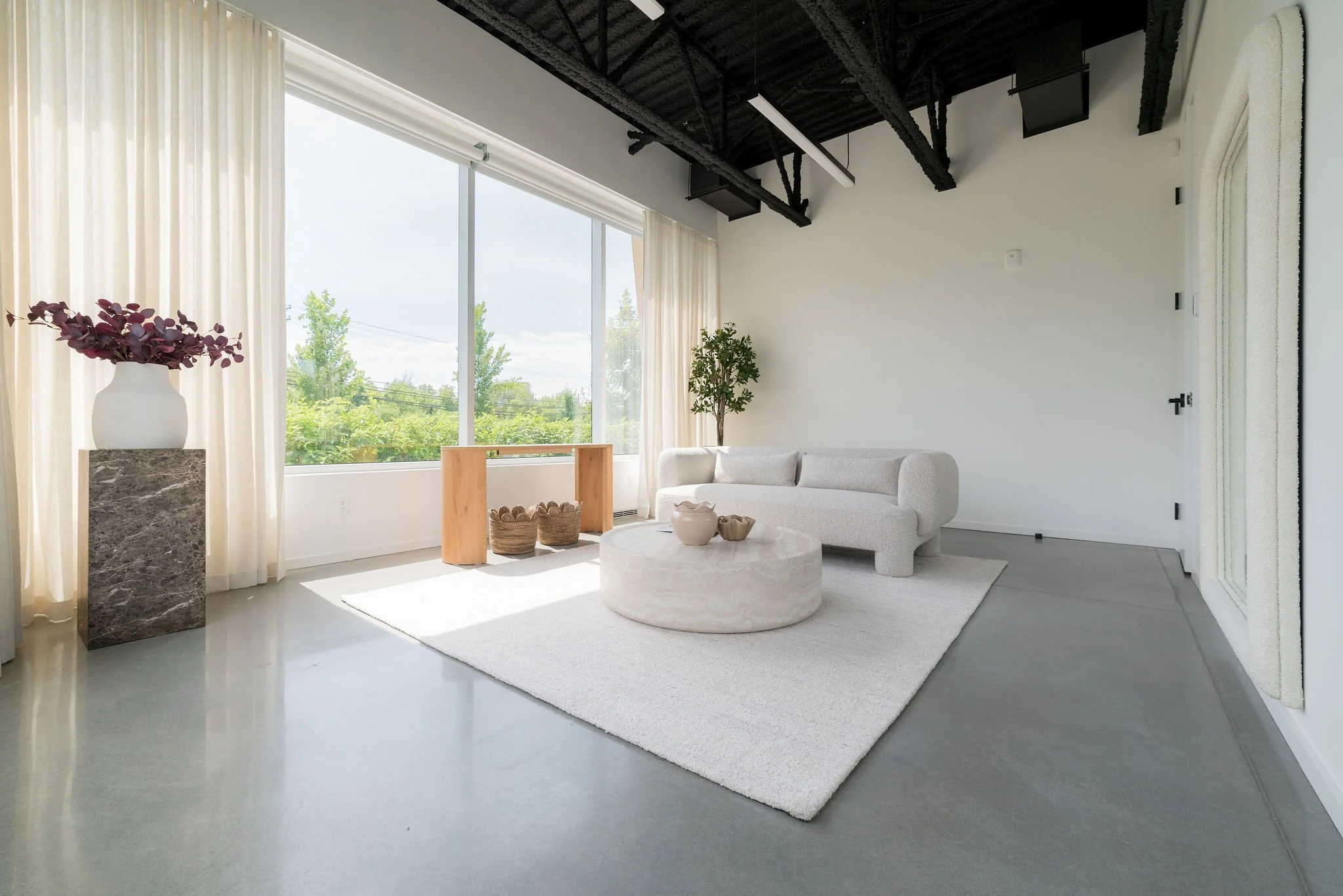Salon moderne avec grande fenêtre laissant entrer beaucoup de lumière naturelle, canapé beige, tapis blanc, table ronde en marbre, décoration minimaliste avec plantes vertes et vases en pierre, plafond noir avec structures apparentes.