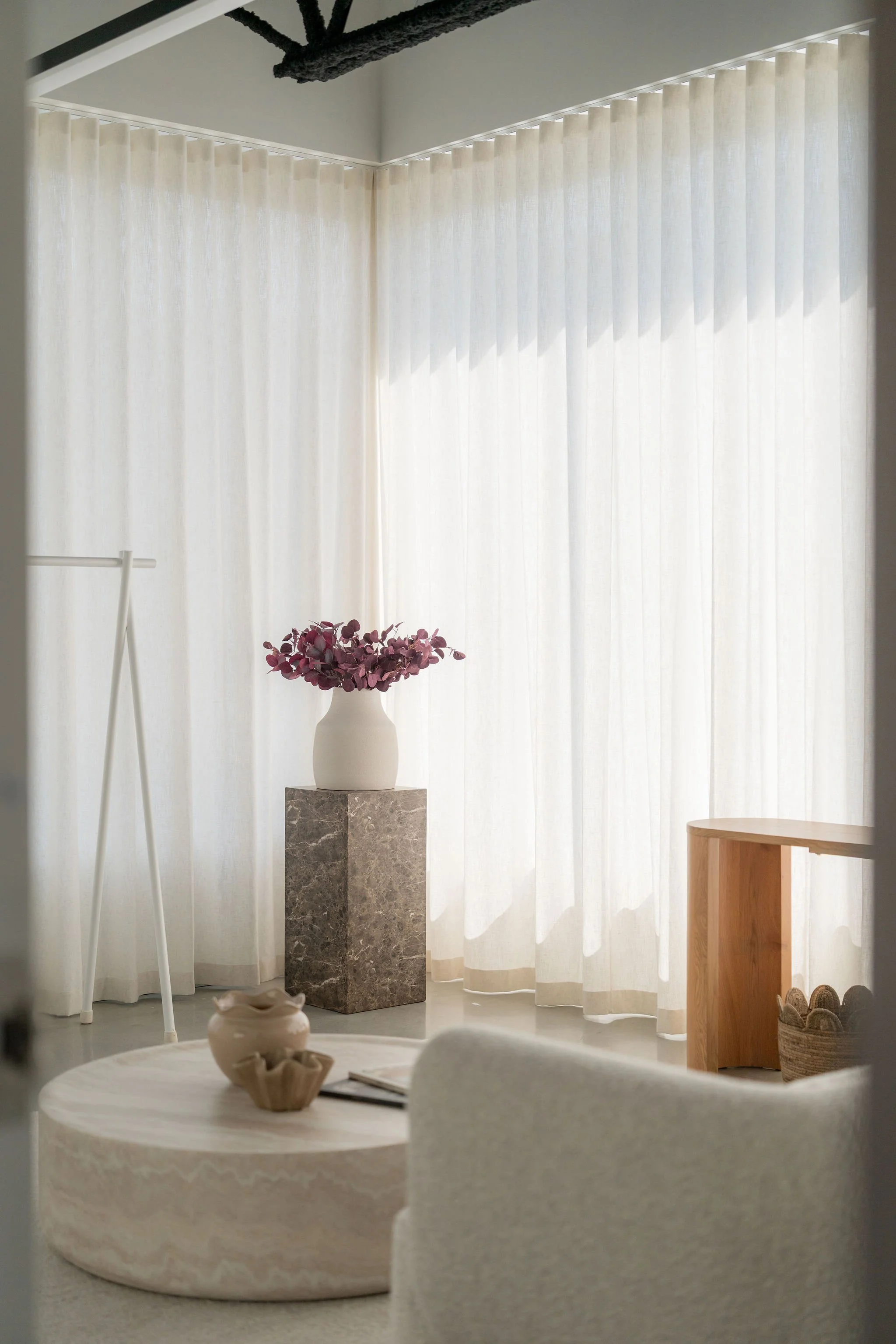 Intérieur minimaliste avec une table ronde en marbre, une vase blanche contenant des fleurs roses, un rideau blanc, et des meubles en bois, lumière naturelle