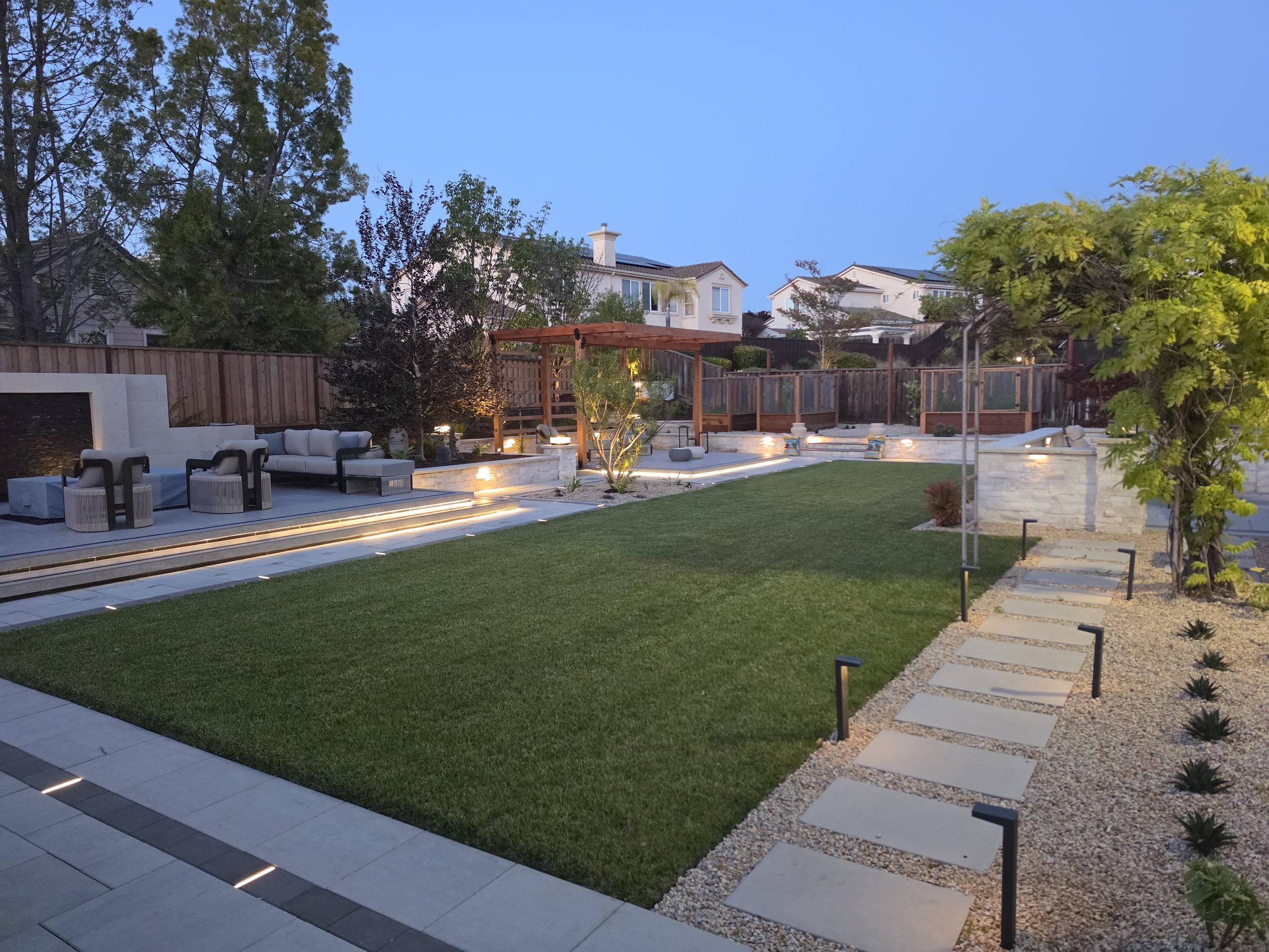 Custom outdoor living space featuring a wood pergola, fire feature, hardscape patio, seating area, landscape lighting, and professionally installed lawn.