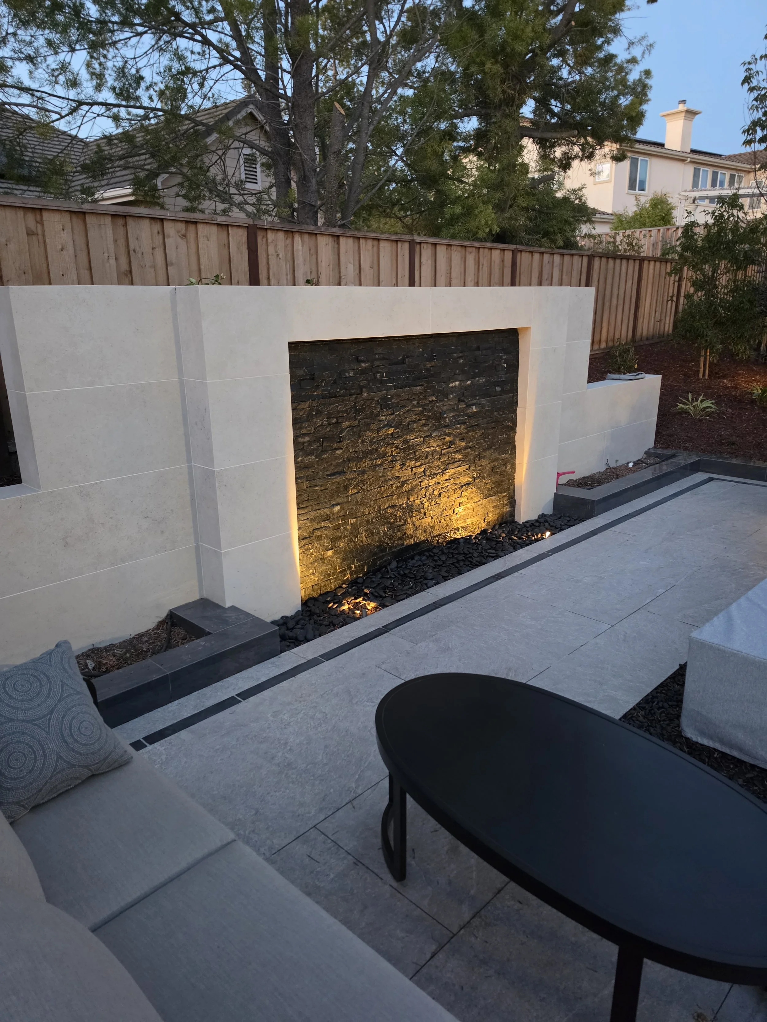 Custom outdoor living space featuring a stone water feature, landscape lighting, hardscape patio, seating area, and professional landscape installation.