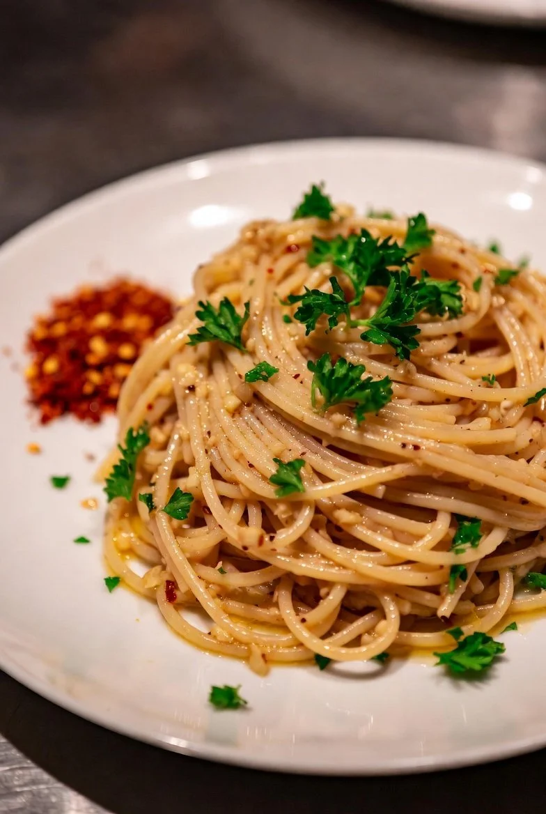 A plate of spaghetti pasta garnished with parsley and red pepper flakes beside a small pile of red pepper flakes.