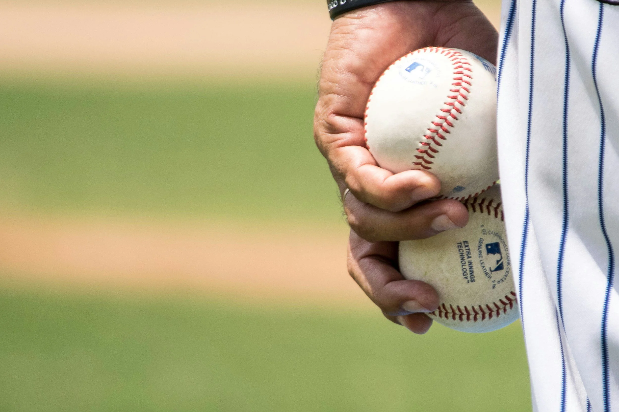 A person wearing a striped sports jersey holding a baseball, with their hand positioned on the ball for a pitch, standing on a field with a blurred grassy background.