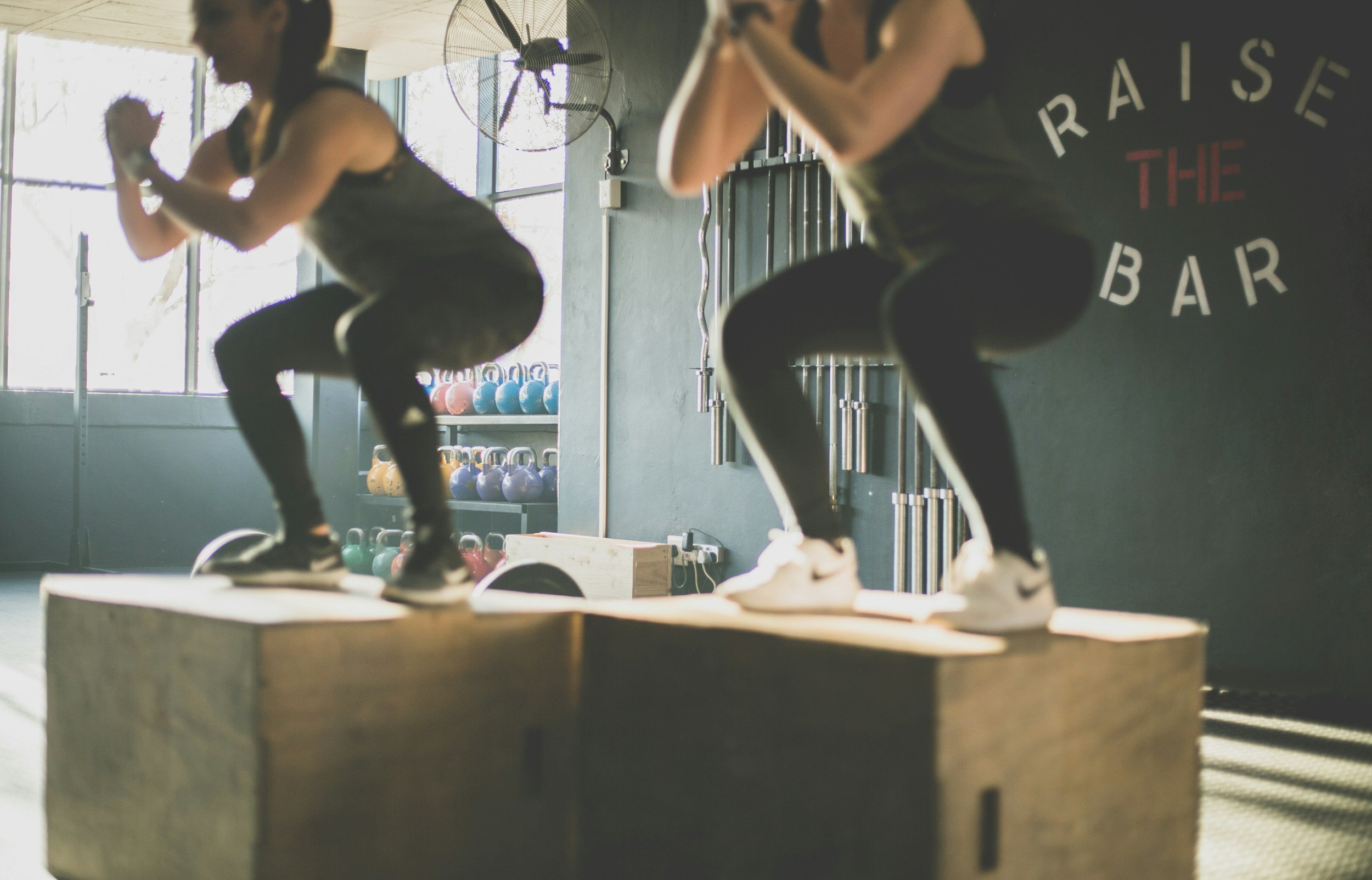 Two women are working out on wooden boxes in a gym, performing box jumps. They are wearing athletic clothing and are focused on their exercise.