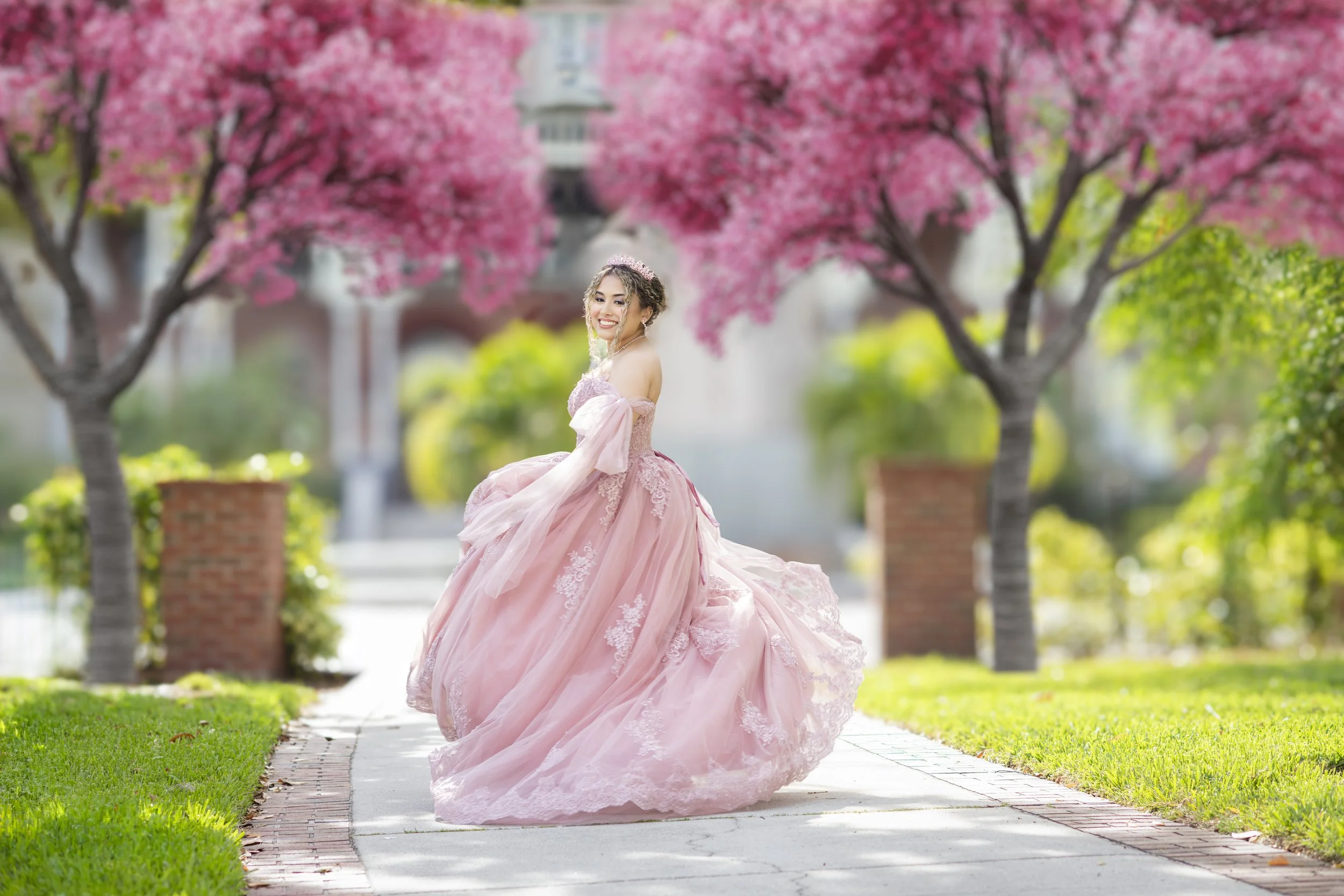 A woman in a pink ball gown with lace and floral details standing on a sidewalk surrounded by pink flowering trees.