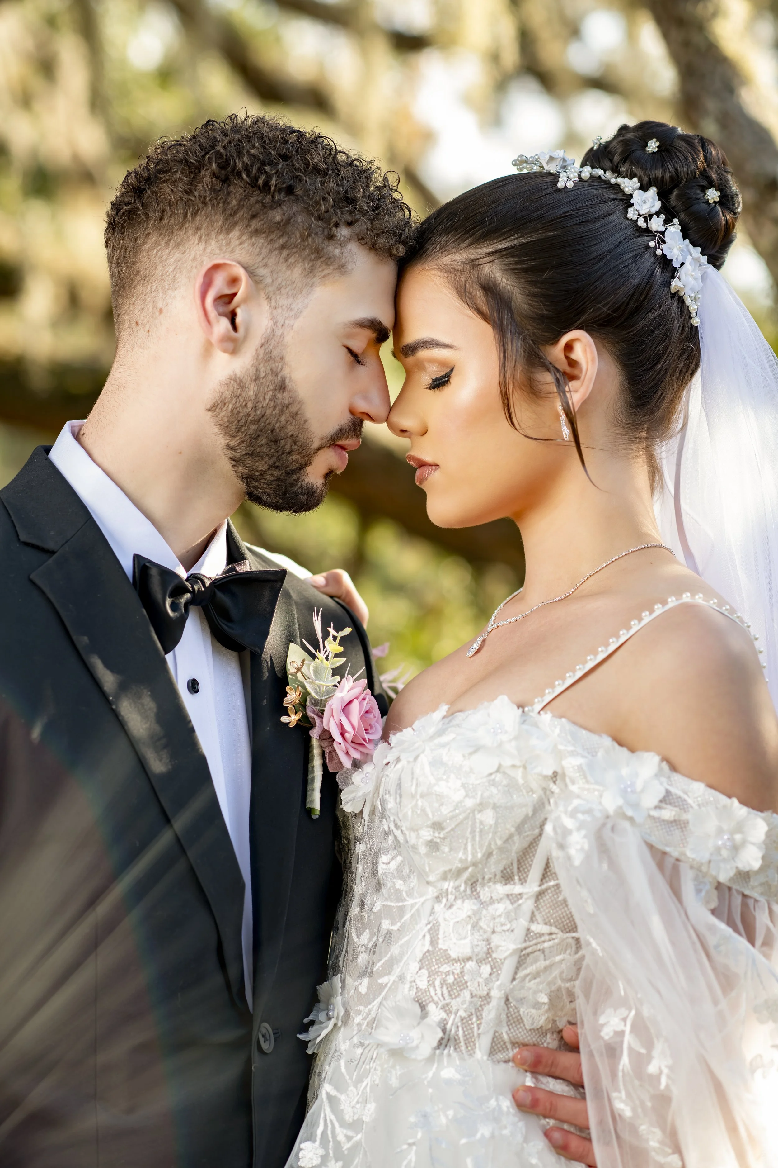 A bride and groom with foreheads touching, eyes closed, outdoors during their wedding, with trees and sunlight in the background.