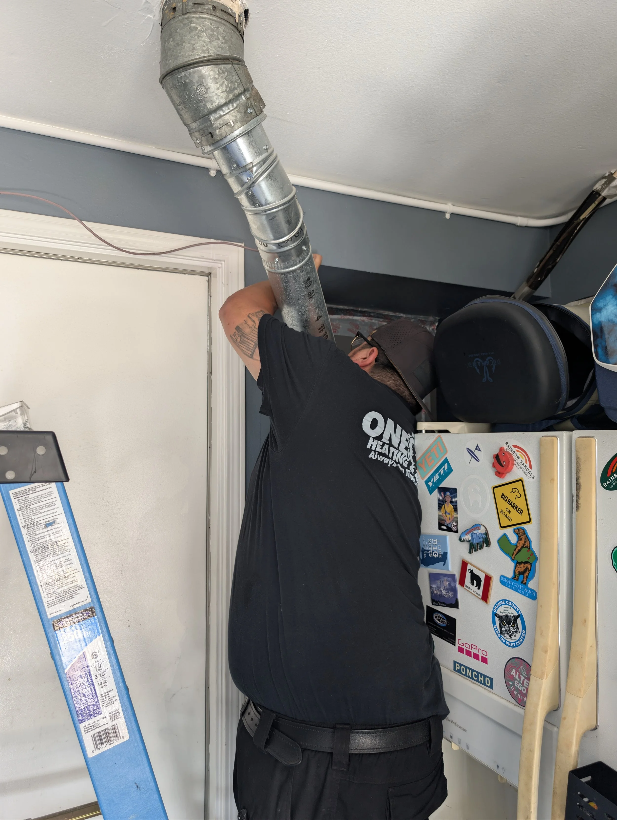 A licensed HVAC technician working on a ceiling duct in a room, with a rainbow-colored sticker visible on a nearby board and various other stickers on a white surface.