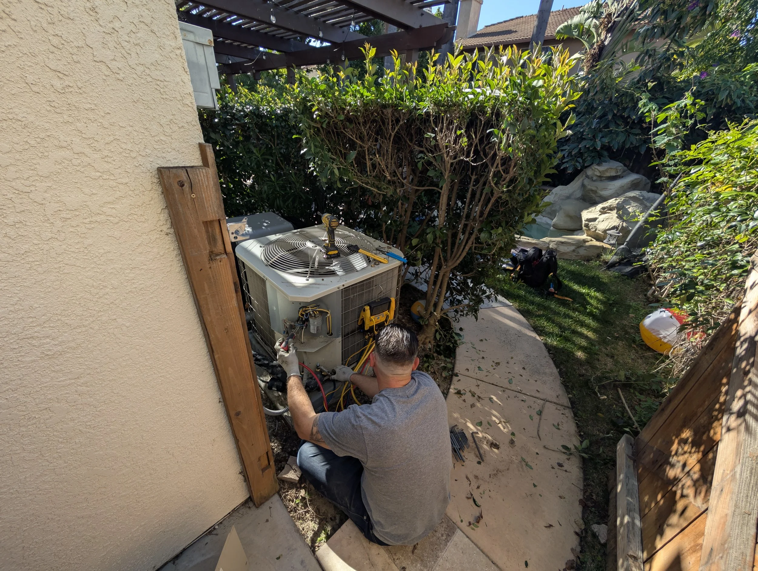 A man working on an air conditioning unit outside a house, surrounded by bushes and a backyard with rocks and plants.