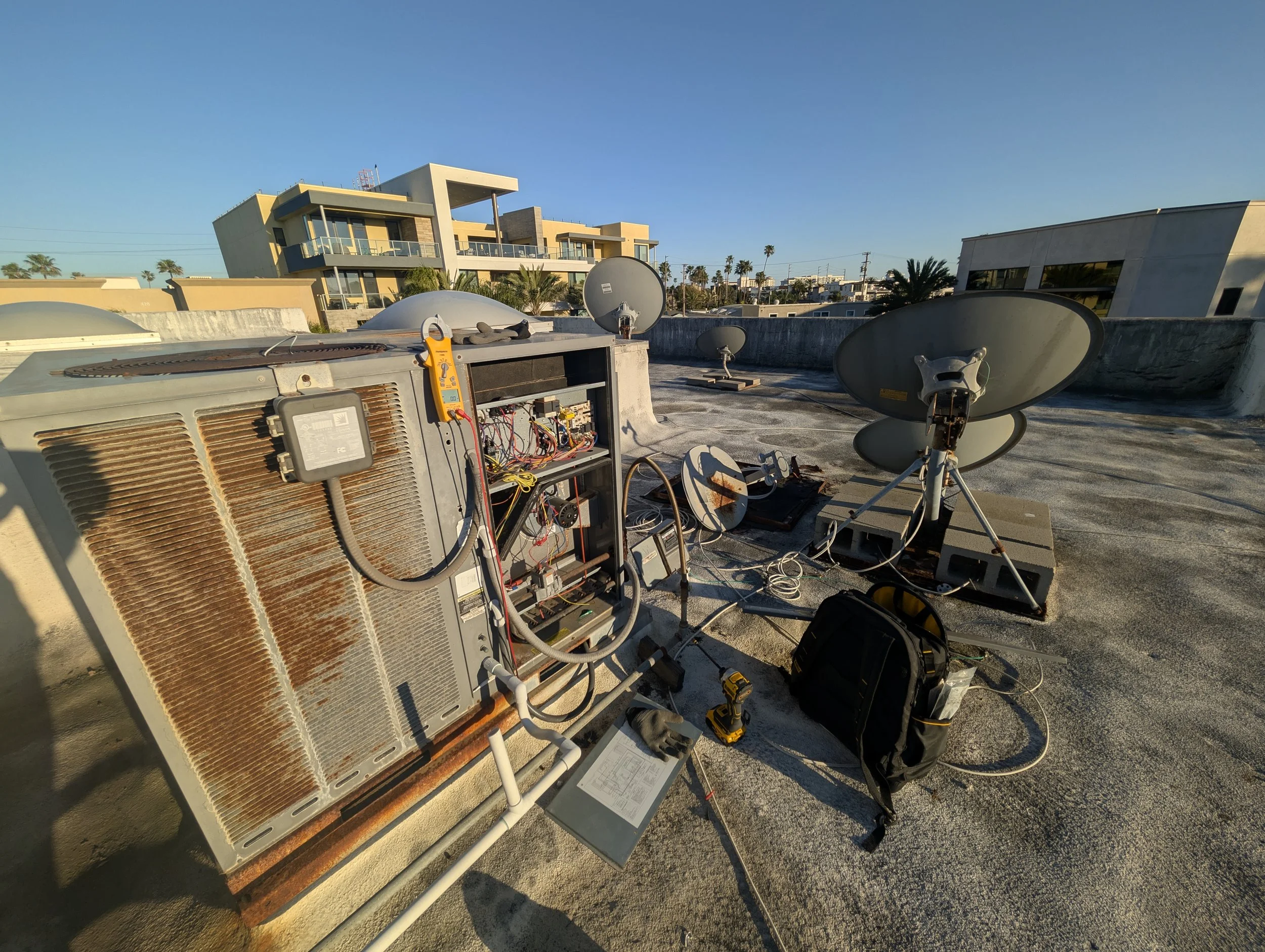 On a building rooftop, an air conditioning unit with a rusted cover and an open panel exposing wiring and components, along with several satellite dishes and equipment, under a clear blue sky.