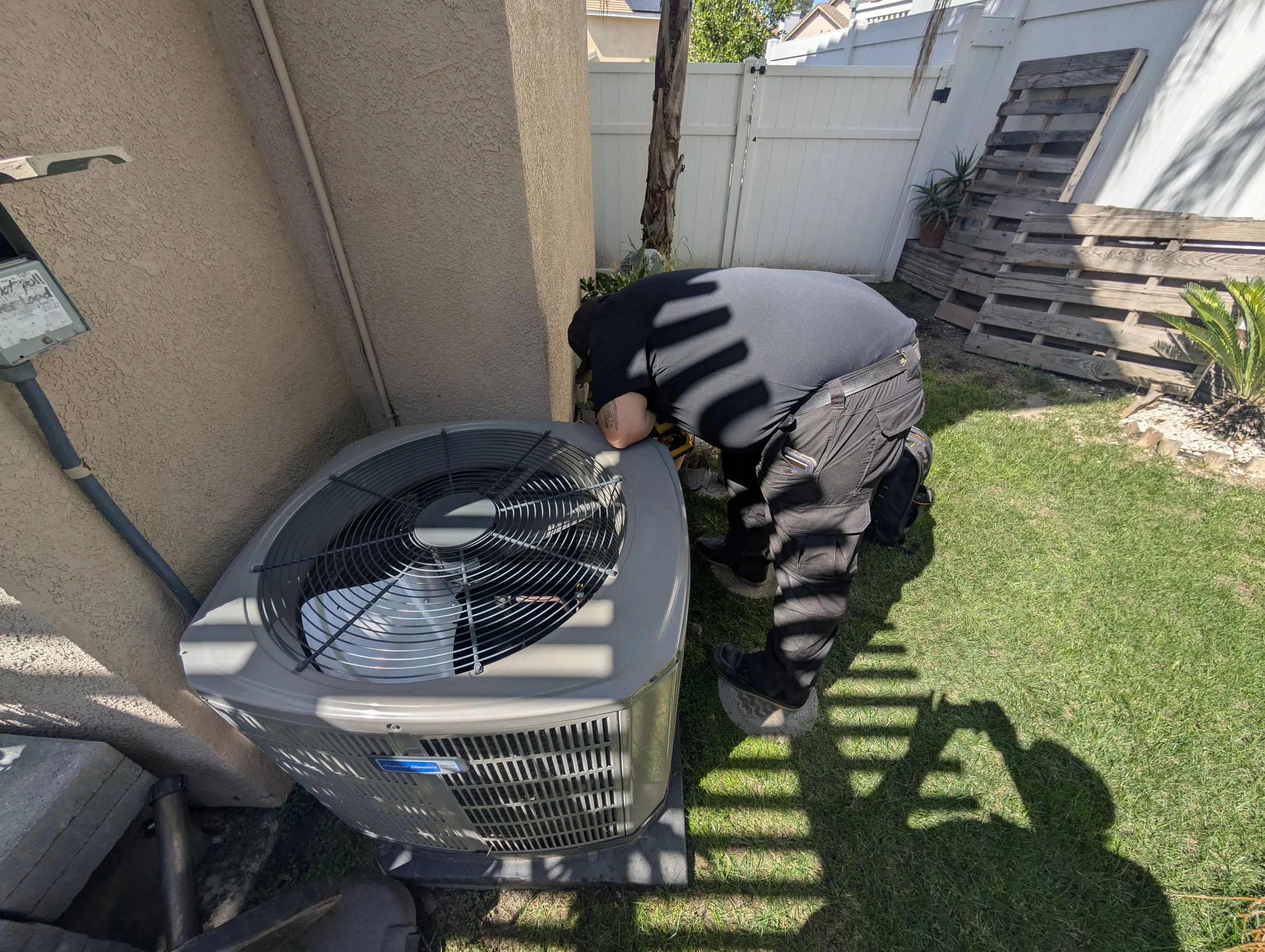 A person wearing a gray shirt and black pants kneels next to an outdoor air conditioning unit, working on it in a backyard with a white fence and some plants.