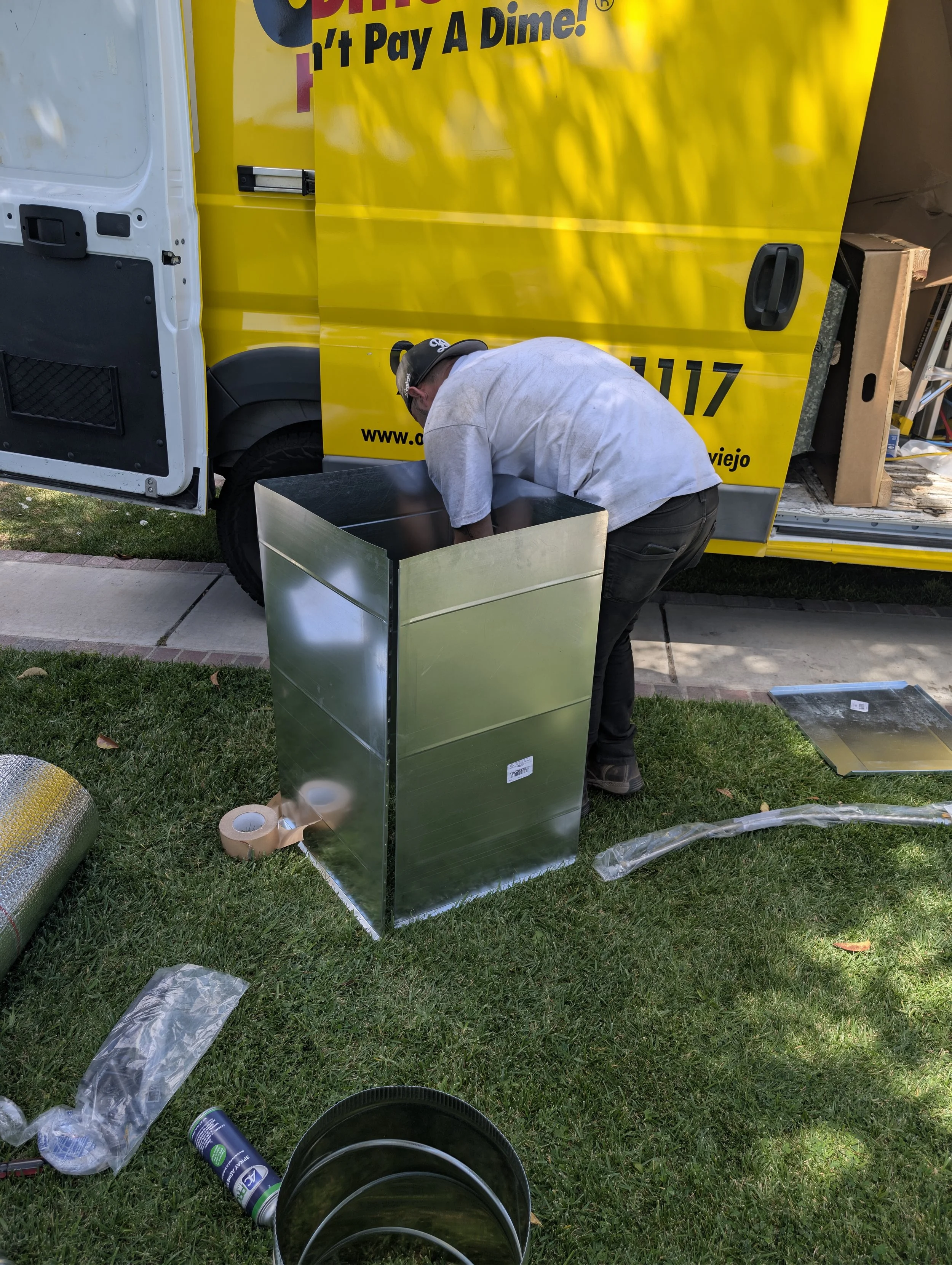 A NATE certified technician is kneeling near a yellow truck with black text and website, working on a large metallic box. There are various tools and materials, including a roll of tape, a spray can, and black buckets, on the grass nearby.