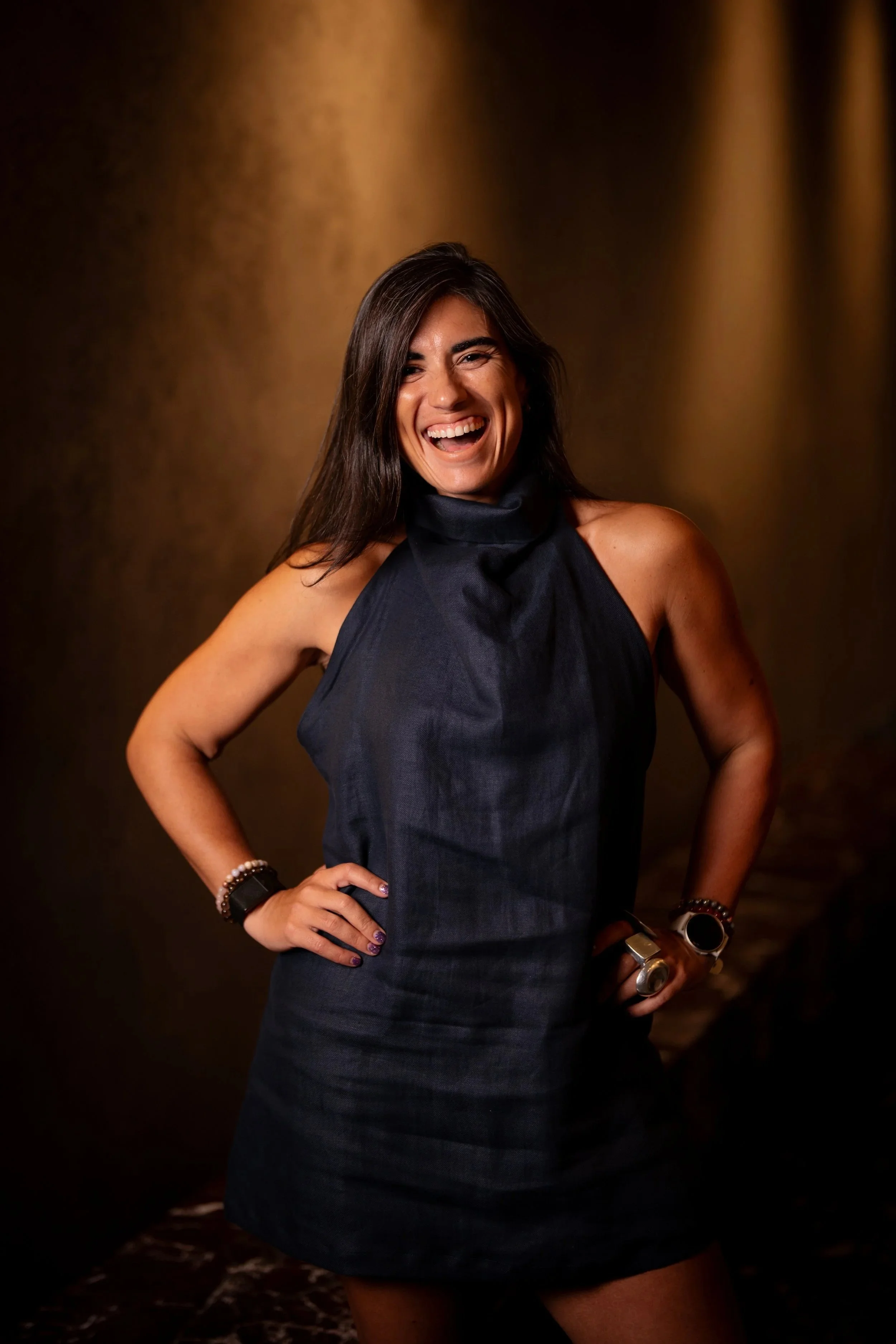Woman with long dark hair smiling, wearing a dark blue dress, standing with hands on hips in a dimly lit room with brown and black background.