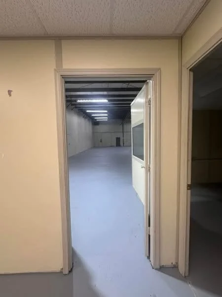 Open door leading into an empty, well-lit warehouse or storage area with exposed ceiling beams and fluorescent lighting.