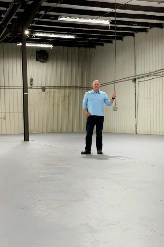 A man standing in an empty warehouse or industrial space, holding a long black pole with a string attached, against beige walls with visible wiring and pipes.