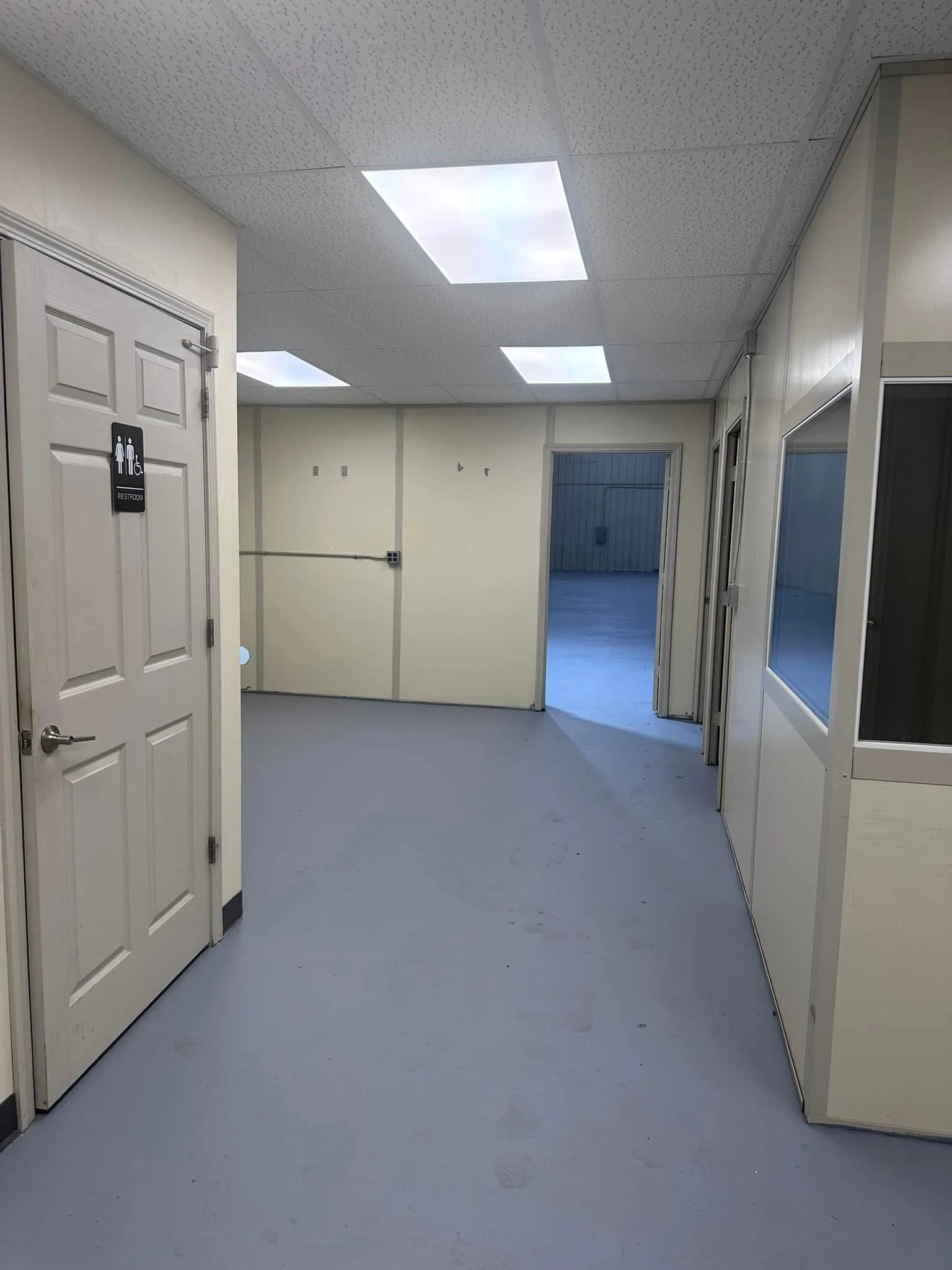 Empty hallway with white walls and ceiling, fluorescent lights, and closed doors, including a restroom sign on the left door.
