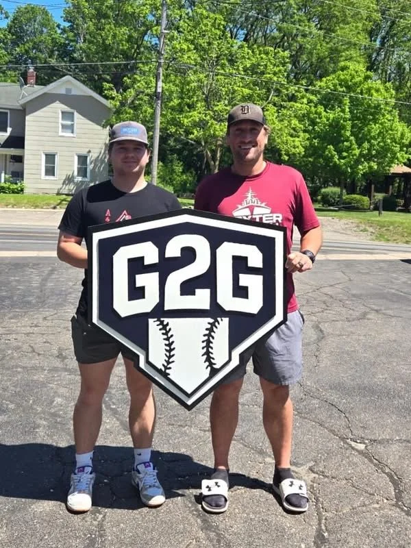 Colin Danek and Hank Dreffs, coaches at G2G, are holding a large sign that reads "G2G" with baseball stitching design.